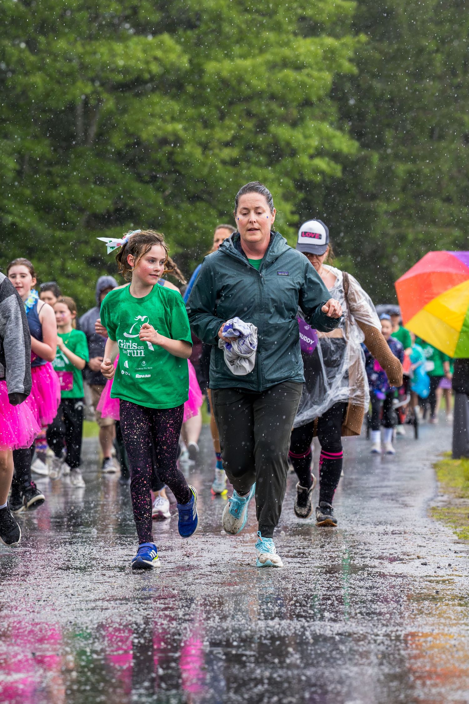 Runners race in the rain during an outdoor charity fun run event, with colorful tutus and umbrellas visible in the background.