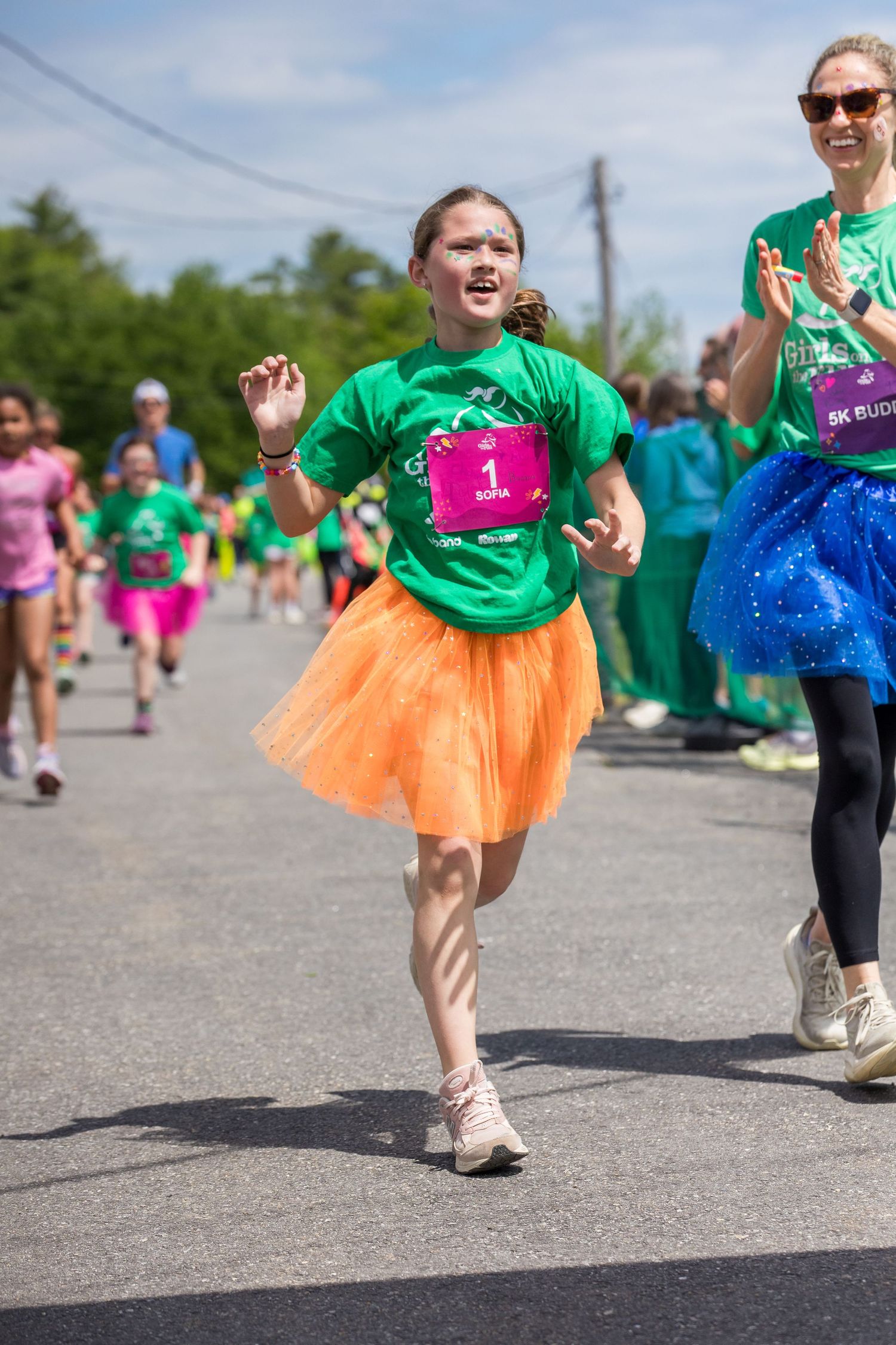 Young girl in green shirt and orange tutu runs in a 5K race, wearing bib number 1, with other participants behind her.