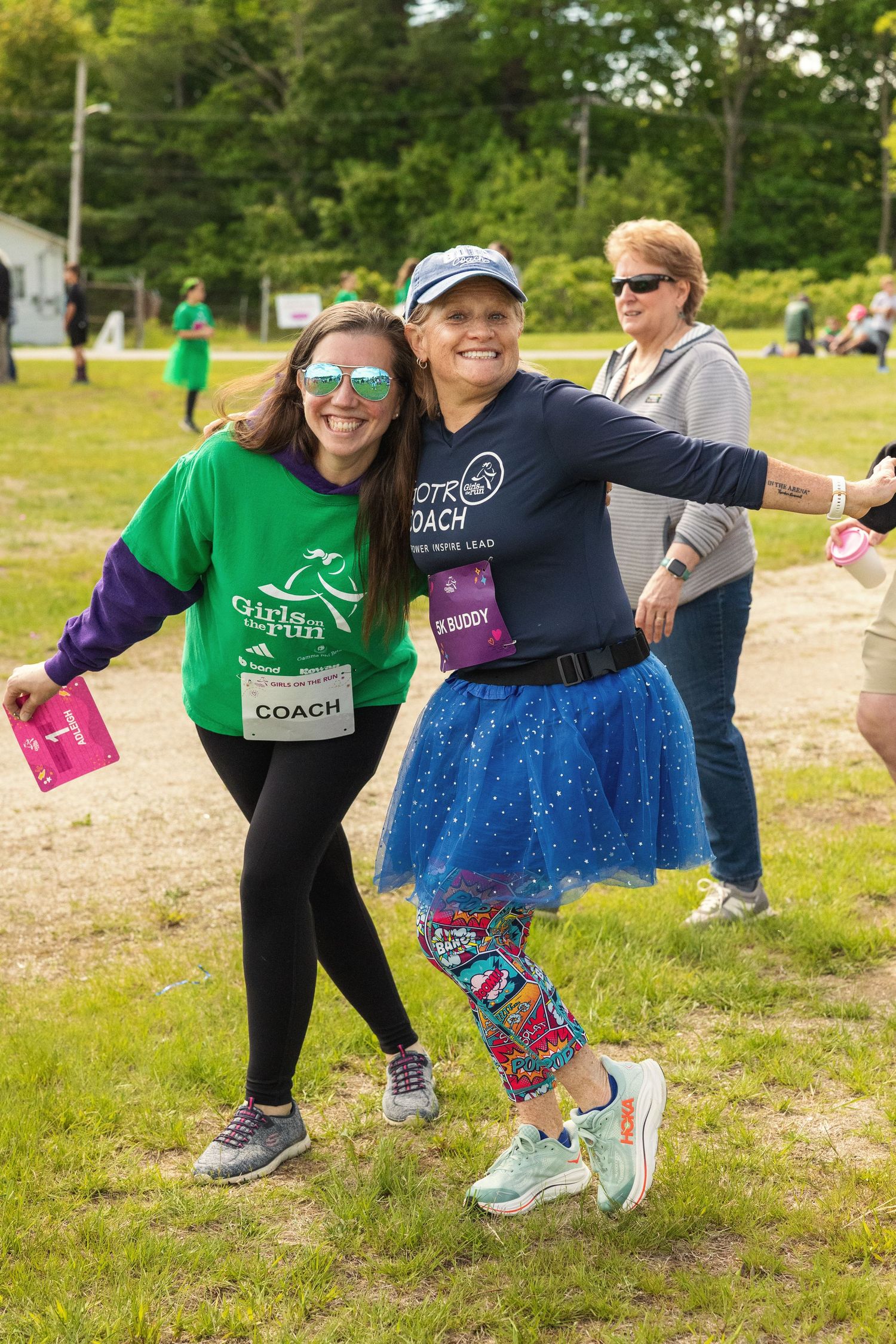 Two women coaches pose together at a Girls on the Run 5K event, wearing race bibs and colorful athletic gear outdoors.
