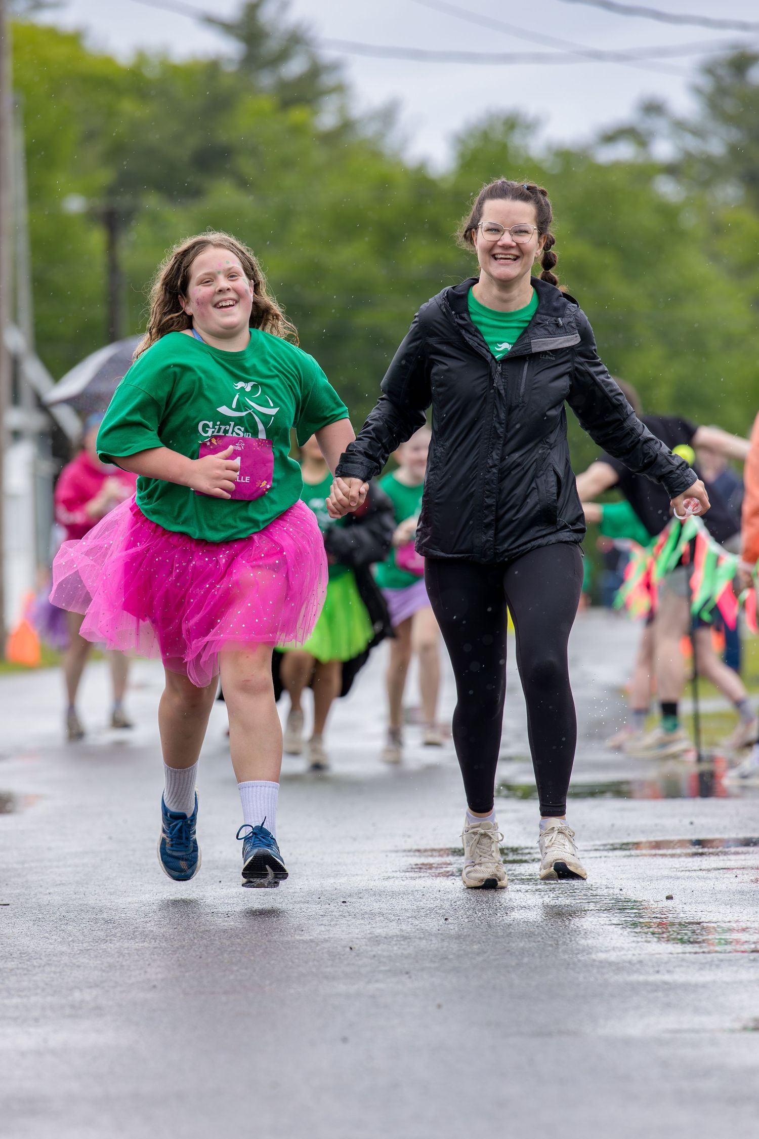 Two smiling women in green shirts and pink tutus joyfully run hand-in-hand during a fun charity race event.