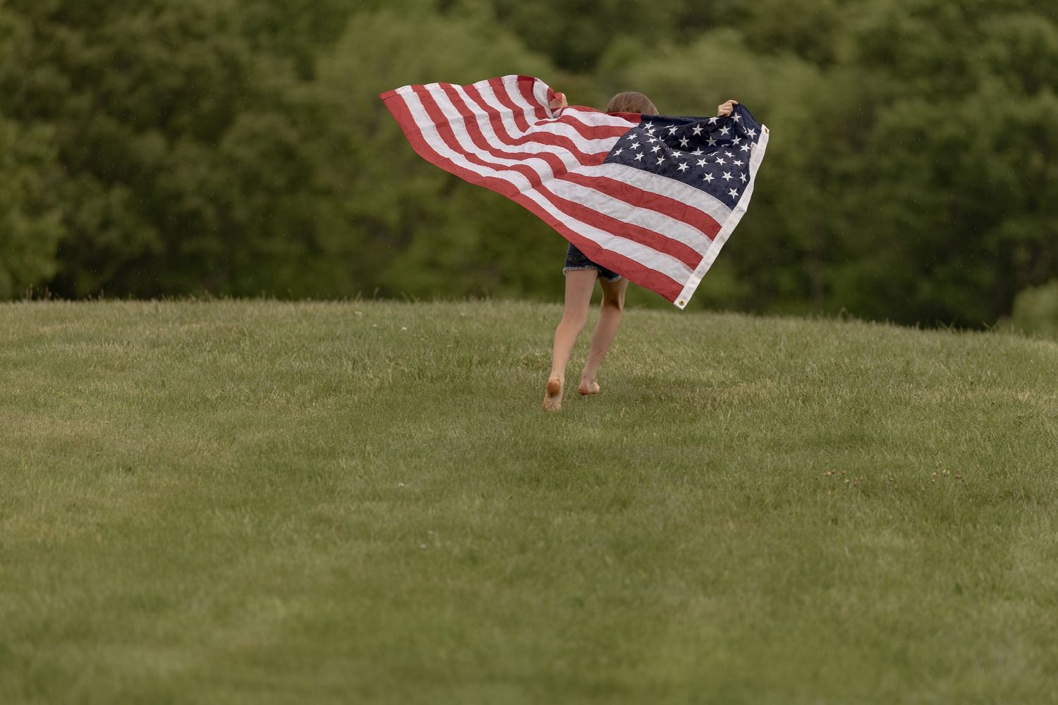 Child running barefoot on green grass while holding a large American flag blowing in the wind.