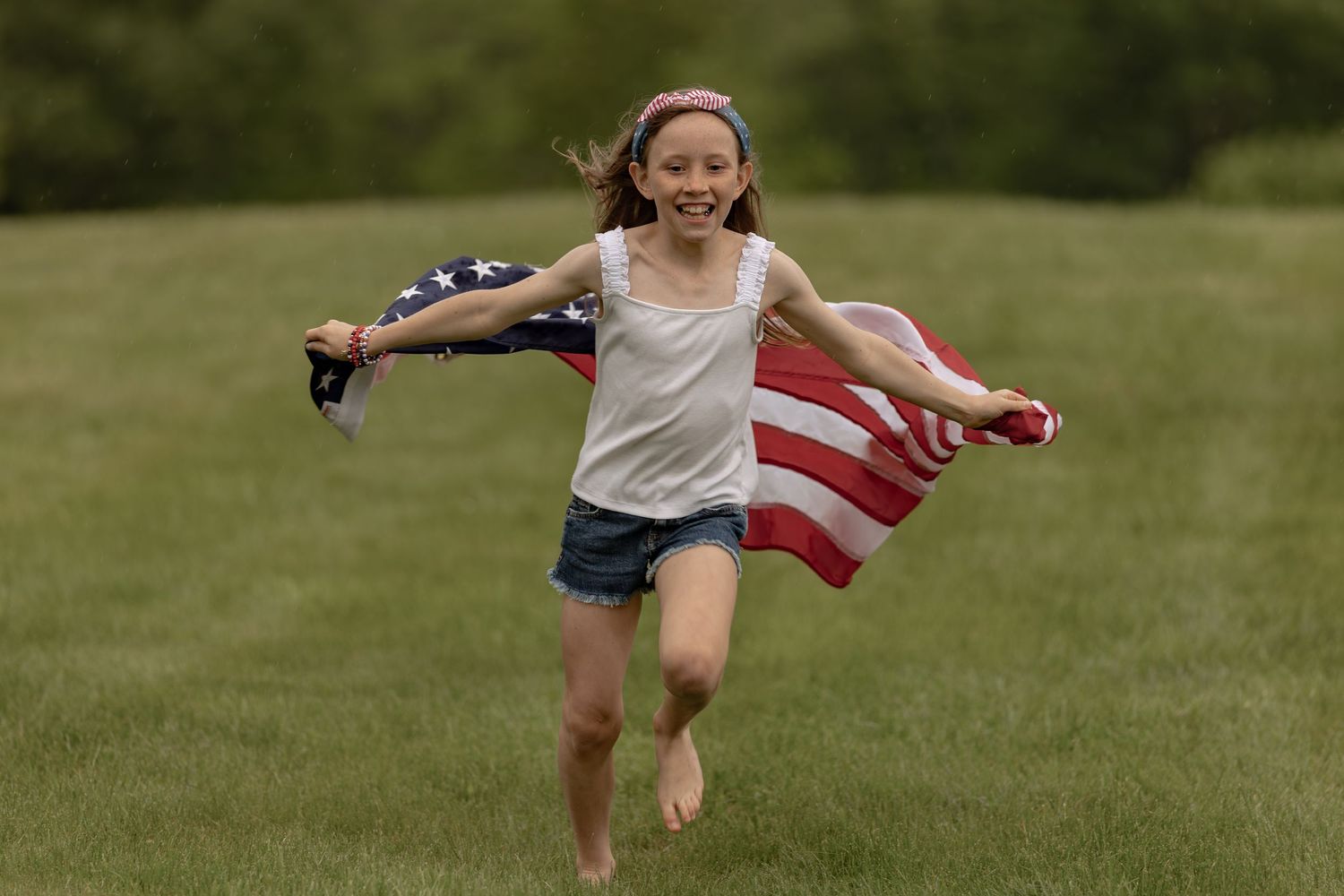 A joyful young girl runs barefoot across a green field, holding an American flag behind her.