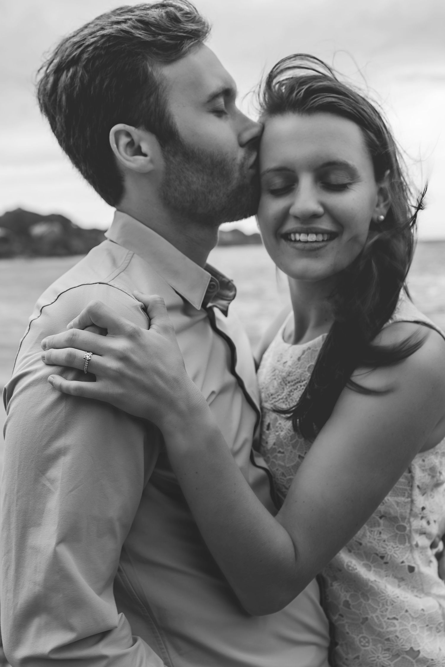Black and white photo of a couple embracing outdoors, man kissing woman on temple as she smiles with eyes closed.