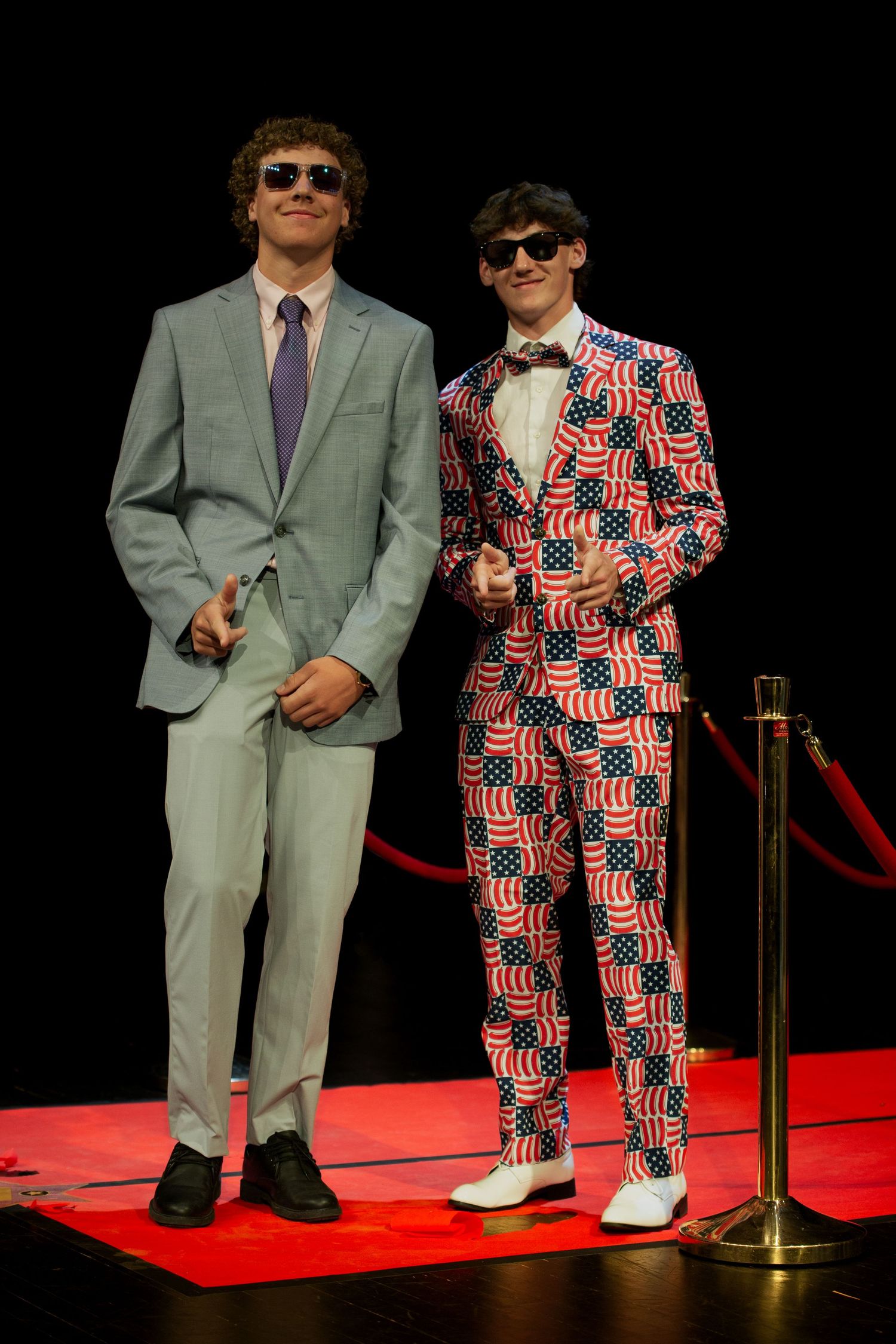 Two young men in suits and sunglasses on red carpet, one in gray suit, one in American flag patterned suit.