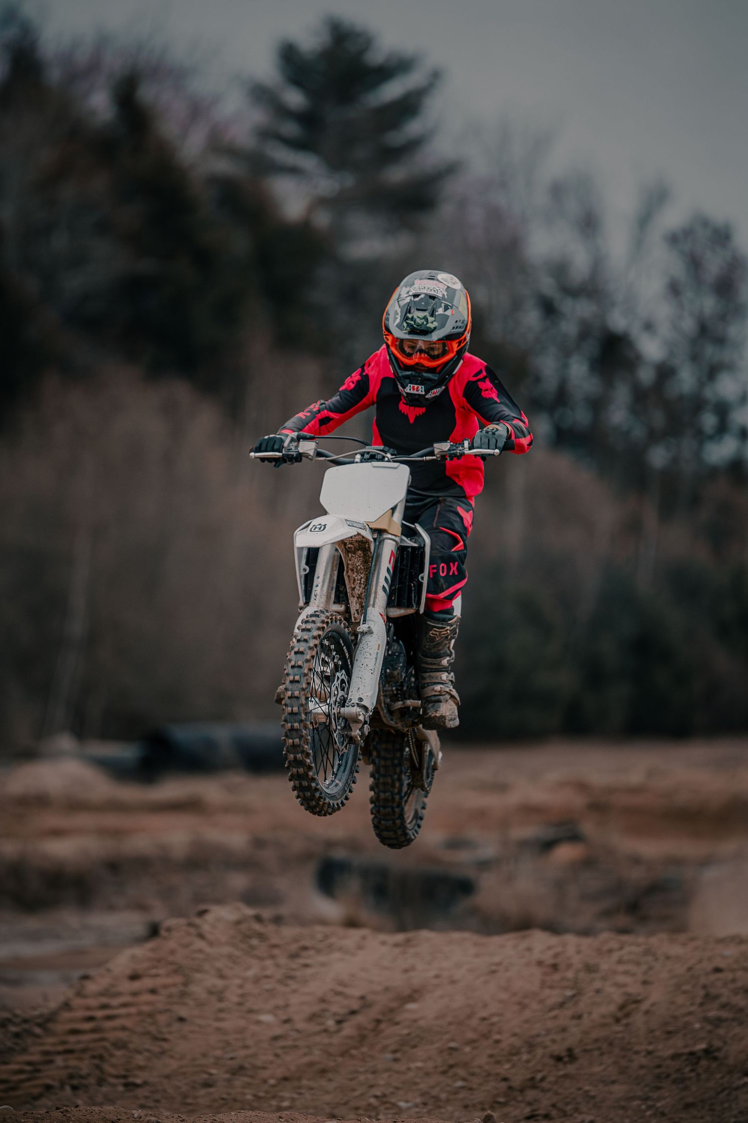 Motocross rider in red gear jumping dirt bike mid-air over sandy terrain with trees in background.