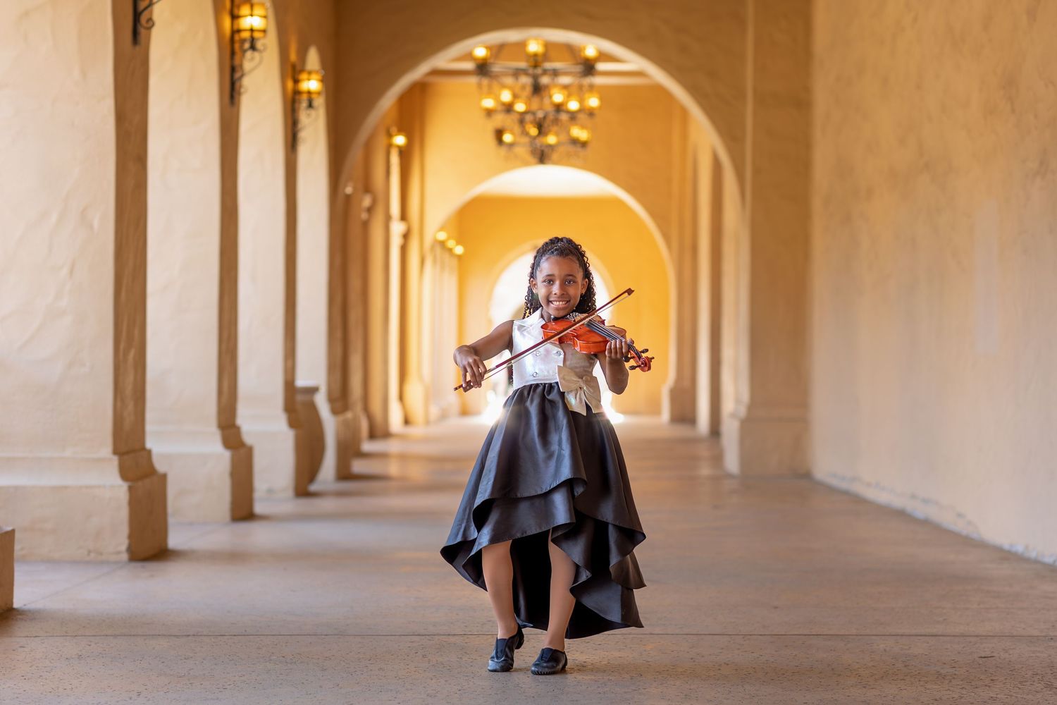 Young girl in black dress plays violin in elegant archway corridor with warm golden lighting.
