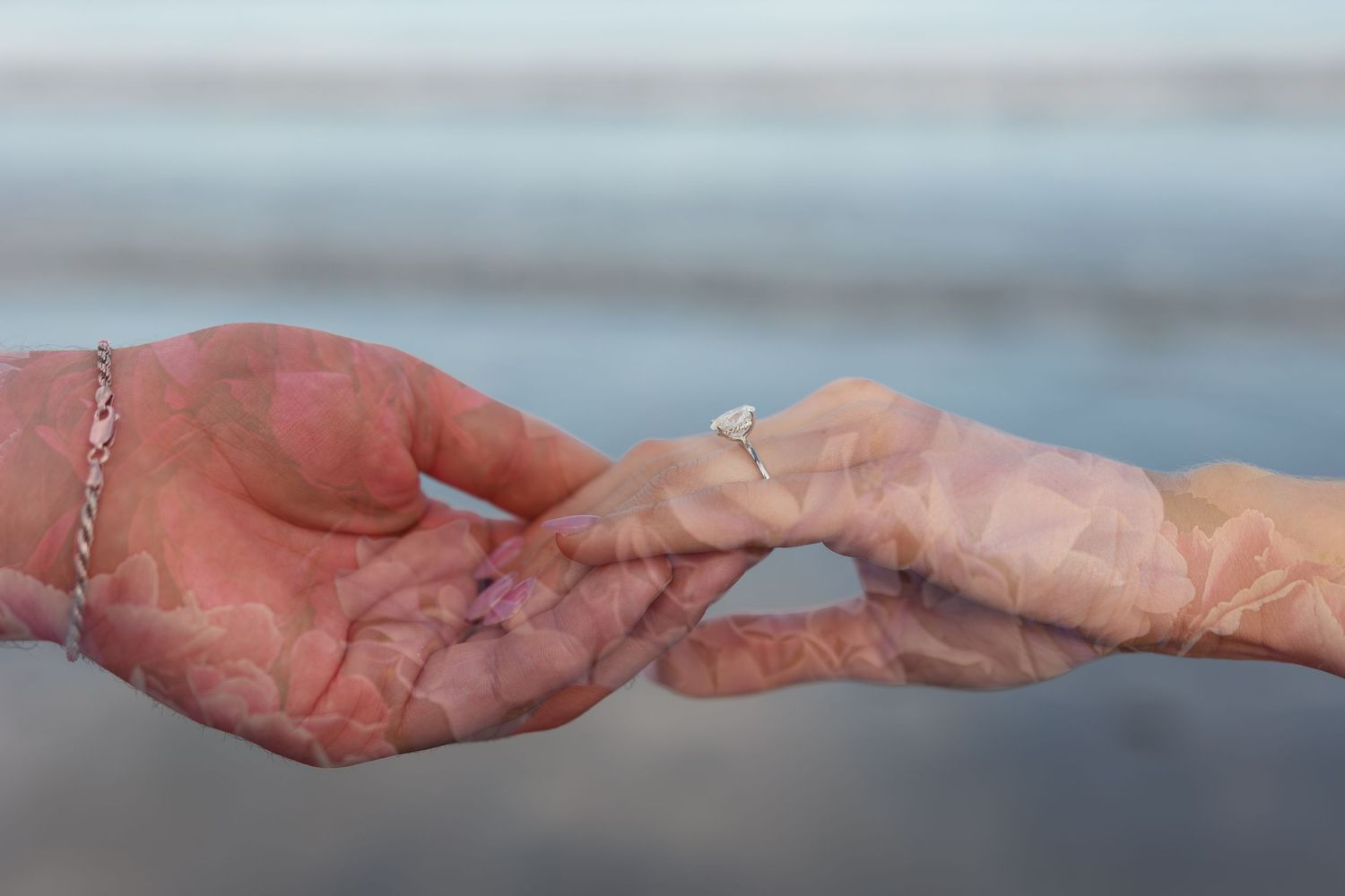 Two elderly hands gently touching, one wearing a diamond ring and bracelet, with a blurred ocean background.