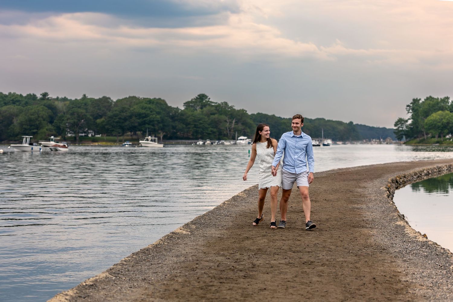 Couple walking hand-in-hand along a sandy waterfront path with boats and lush trees in the background at dusk.