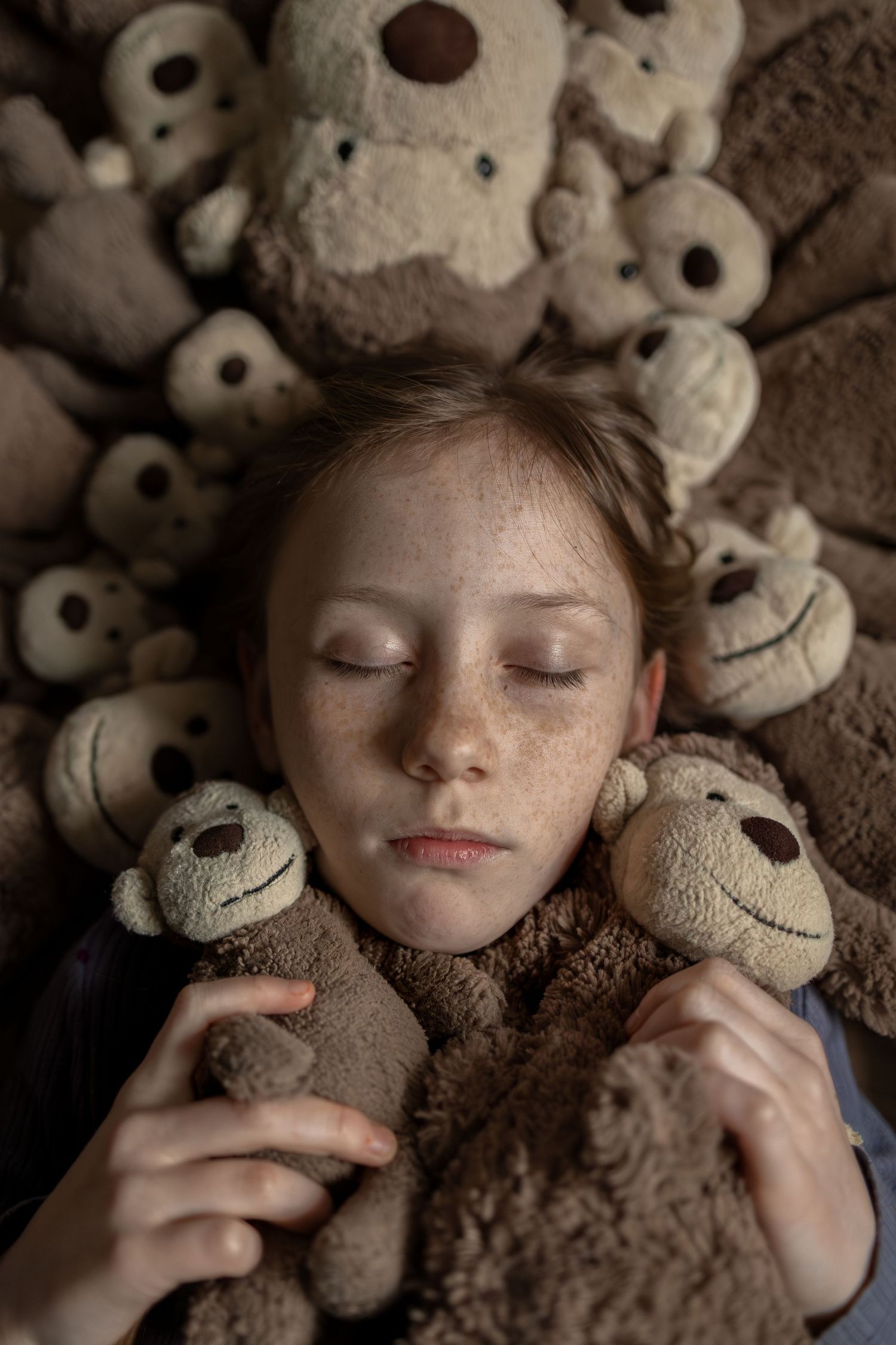 A freckled girl with closed eyes rests peacefully surrounded by numerous brown teddy bears and stuffed animals.