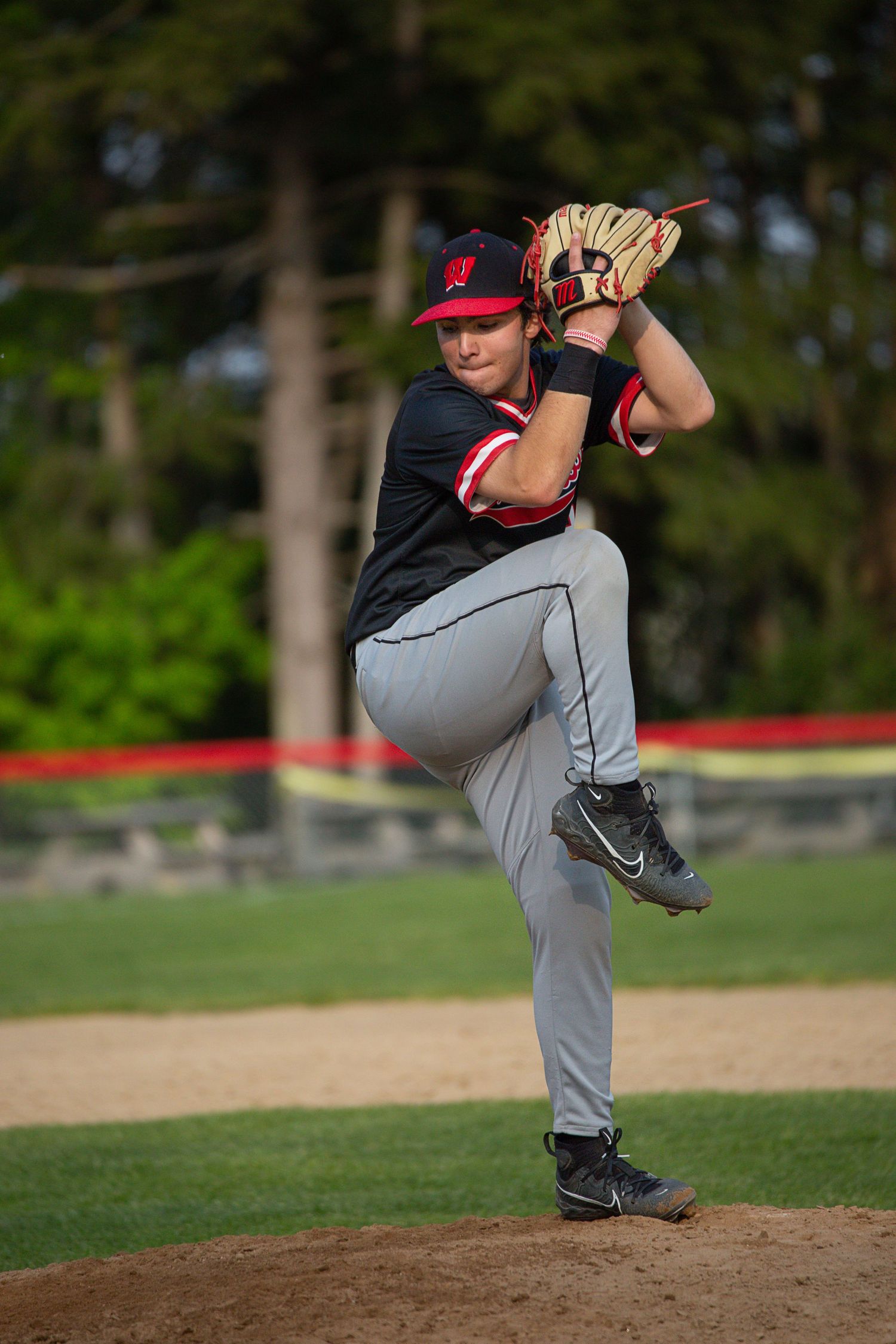 Young baseball pitcher in black and red uniform winds up to throw on the mound during a daytime game.
