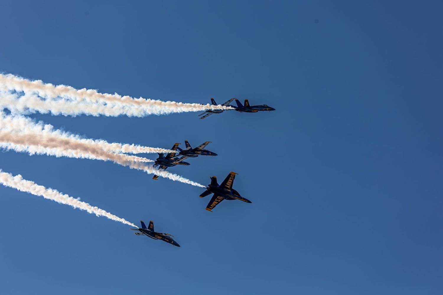 Blue Angels F/A-18 fighter jets perform aerial maneuvers leaving white smoke trails against bright blue sky.