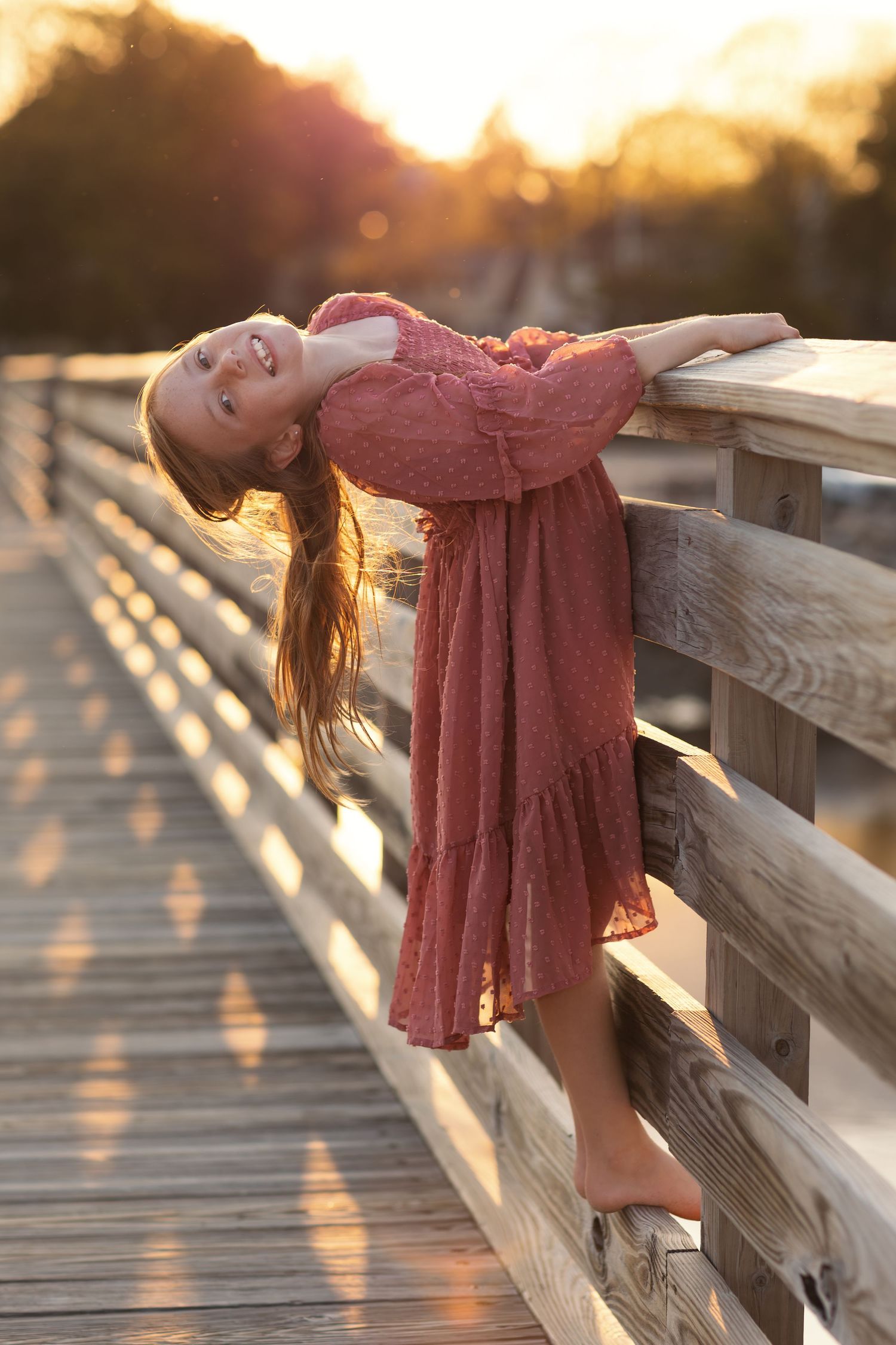 Child in flowing dress leans back on wooden boardwalk railing during golden hour.