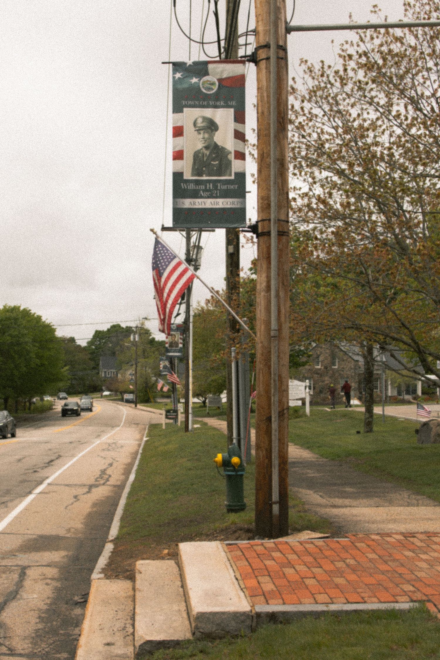 A small-town street lined with American flags and a military memorial banner on a utility pole beside a brick sidewalk.
