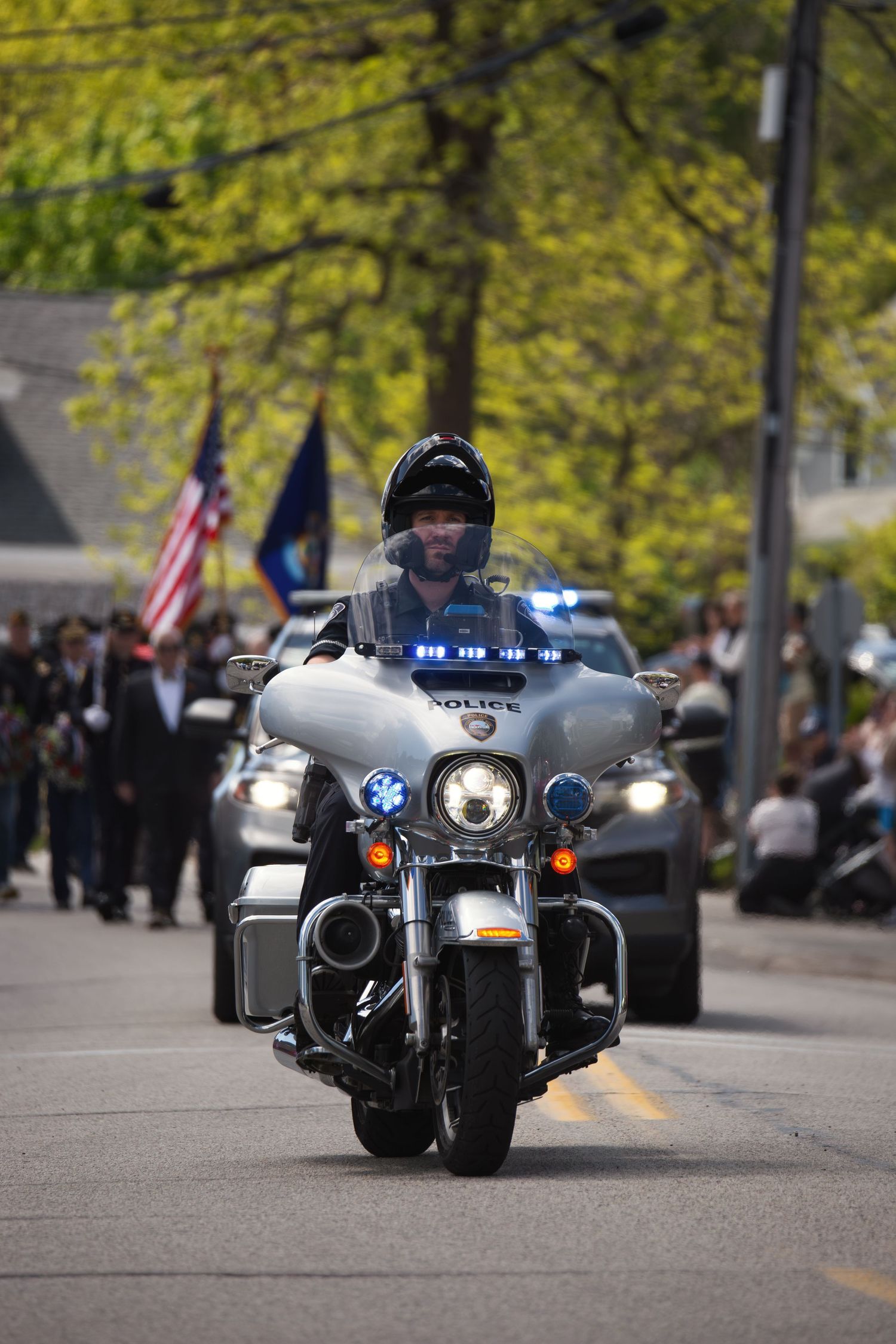 Police officer riding a motorcycle leads a parade down a tree-lined street with flags and vehicles following behind.