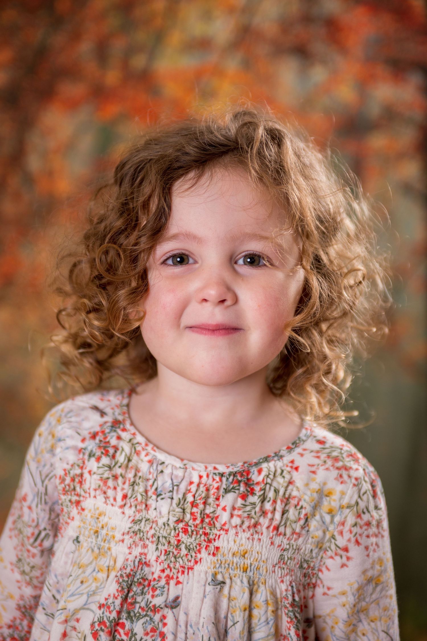 Curly-haired toddler girl in floral dress smiling softly against autumn bokeh background.