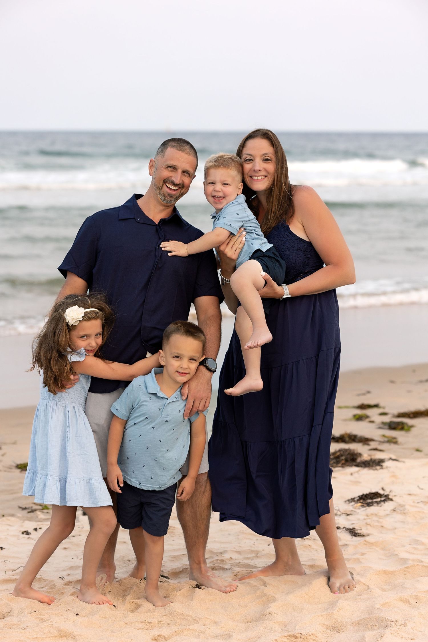 A family of five wearing coordinated navy and blue outfits poses together on a sunny beach with ocean waves in background.