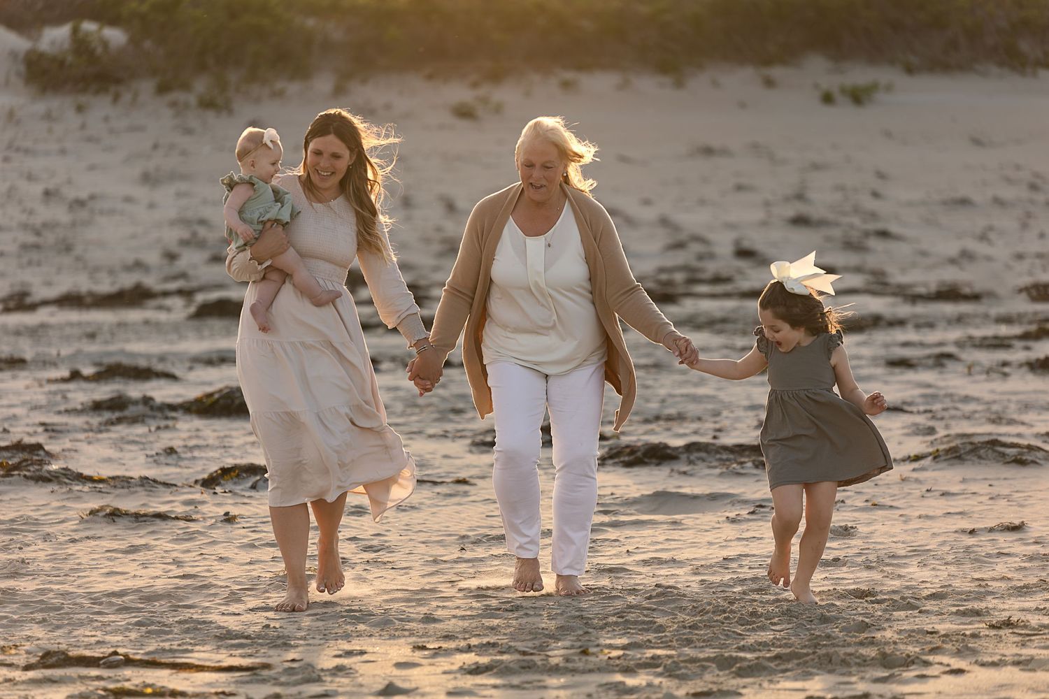 Three generations walking together on a sandy beach at sunset in light colored clothing.