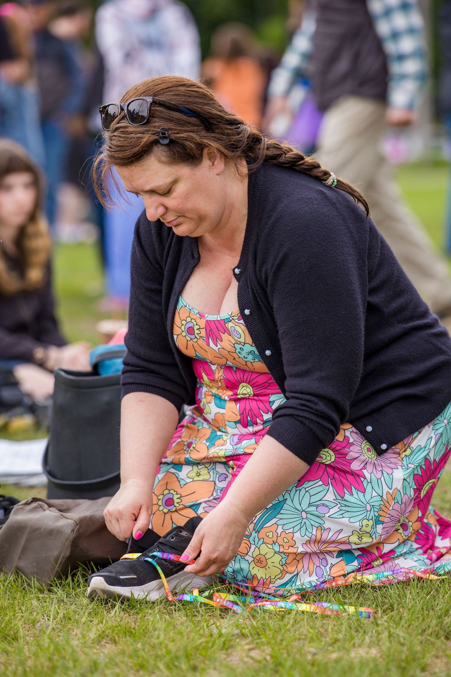 Woman in floral dress sitting on grass tying colorful shoelaces at an outdoor event.
