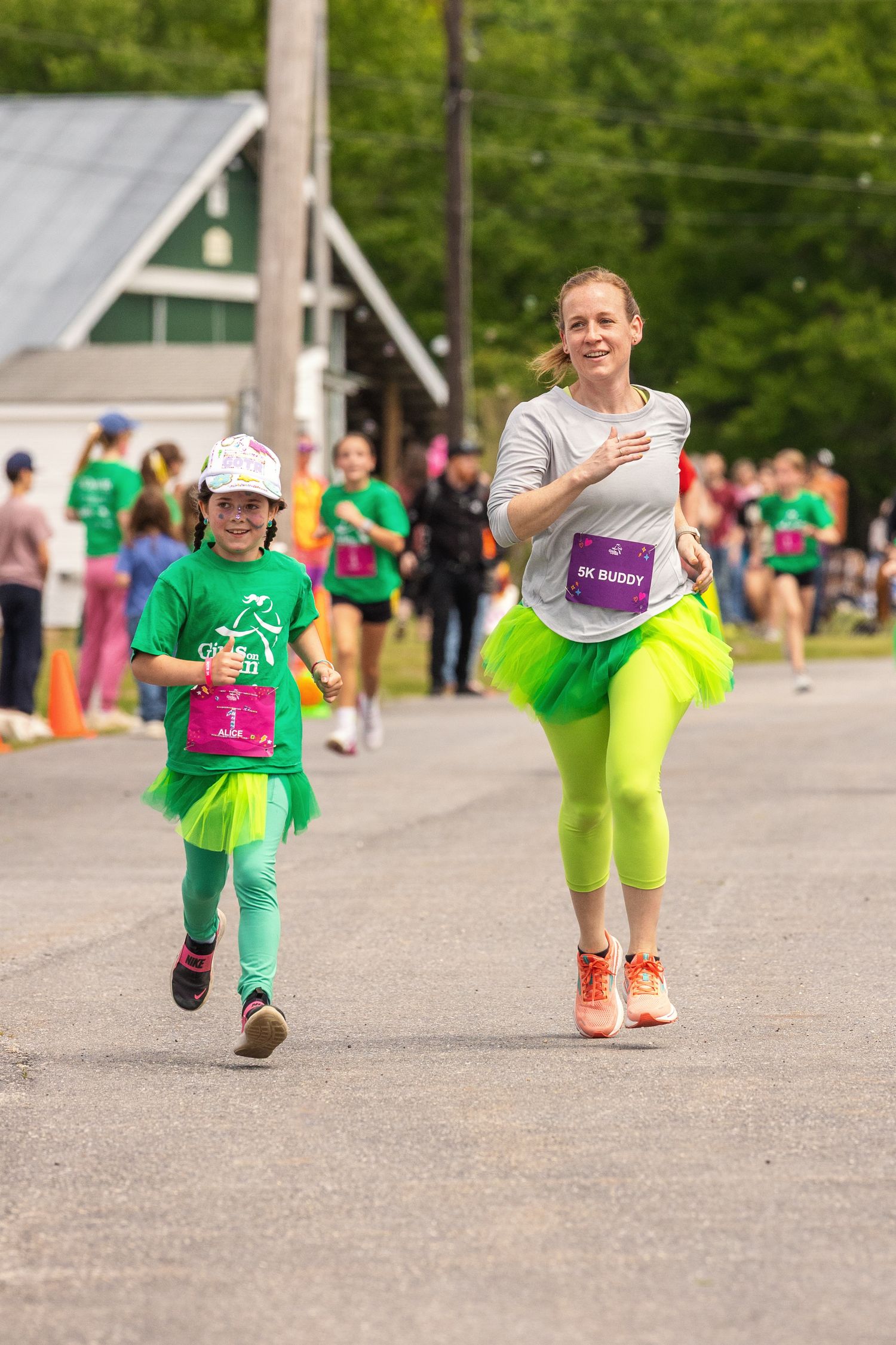 Woman and young girl in neon green tutus running together in a 5K buddy race on a paved path.