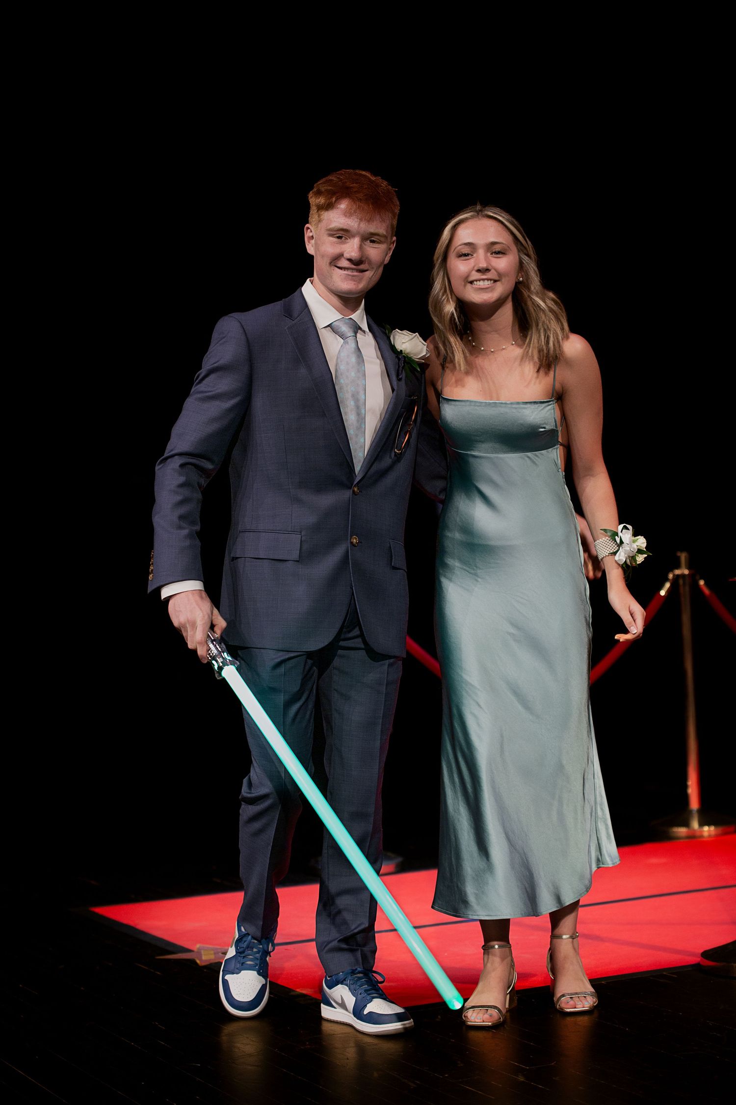 Teen couple posing on red carpet at prom, boy in navy suit with lightsaber, girl in sage satin dress.