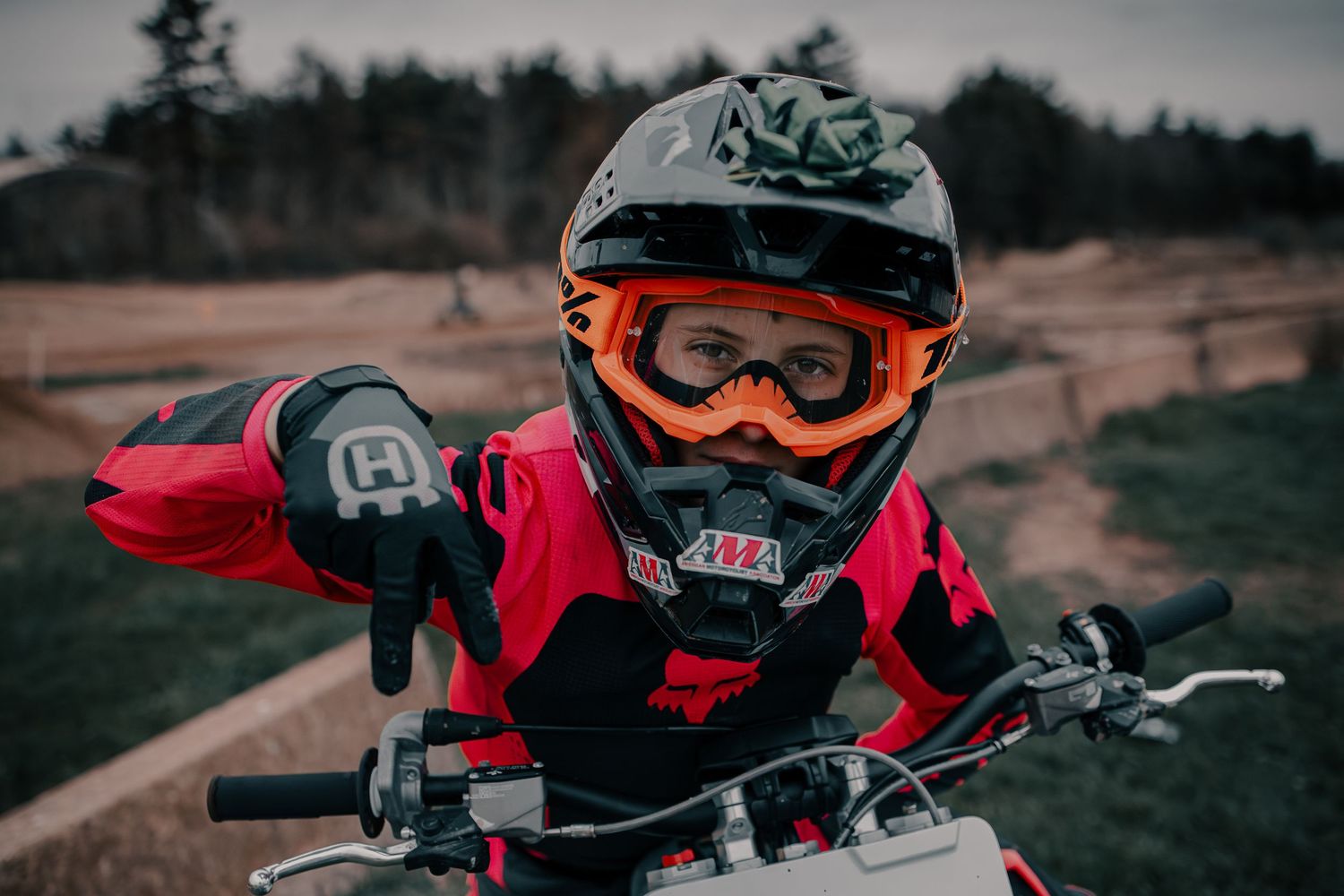 Young motocross rider wearing red gear, orange goggles, and black helmet poses on a dirt bike at an outdoor track.