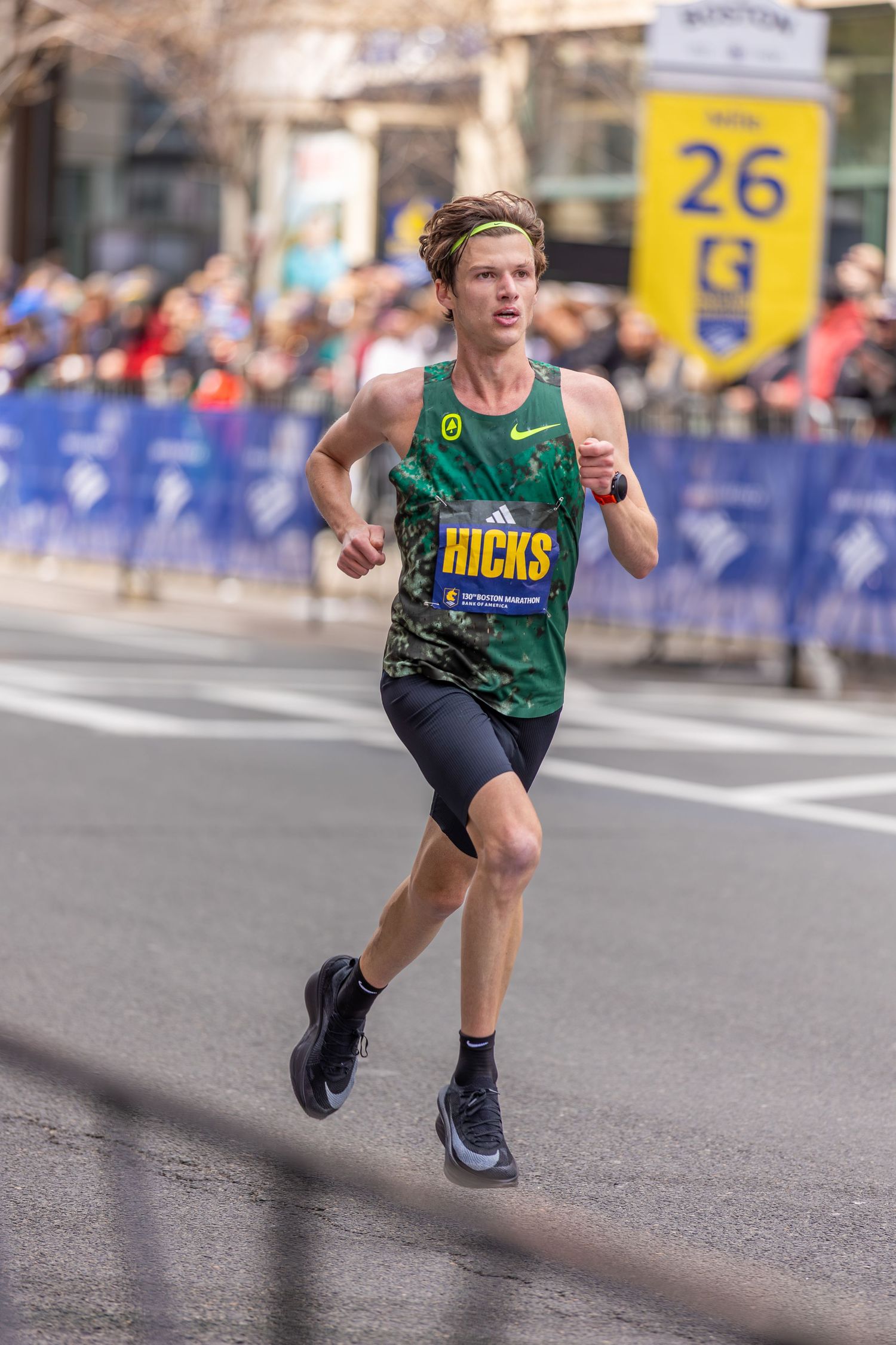 Runner wearing a green Hicks Nike jersey competes at mile 26 of the Boston Marathon on a city street.