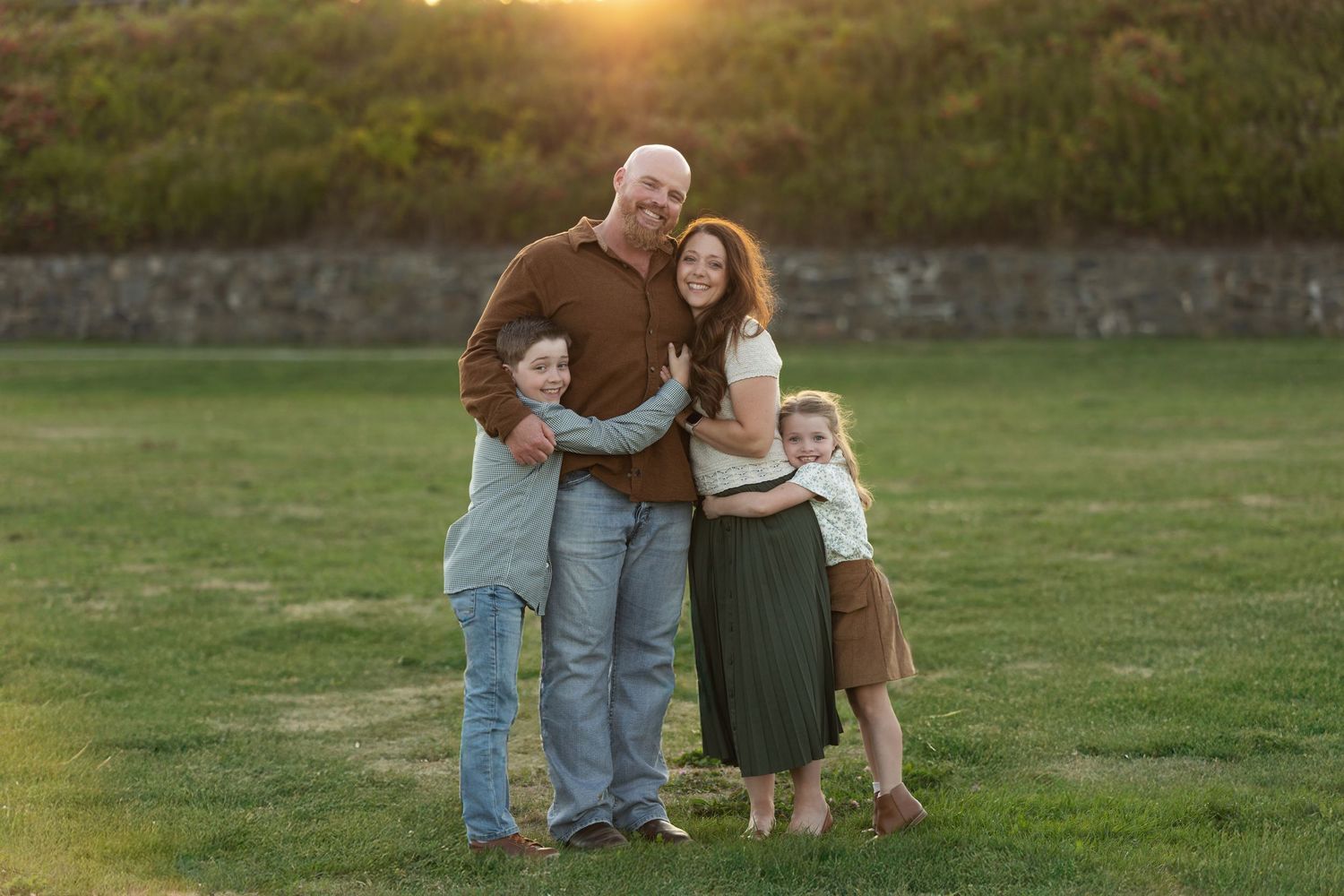 Family of four posing together on green grass at golden hour sunset, parents hugging two young children outdoors.