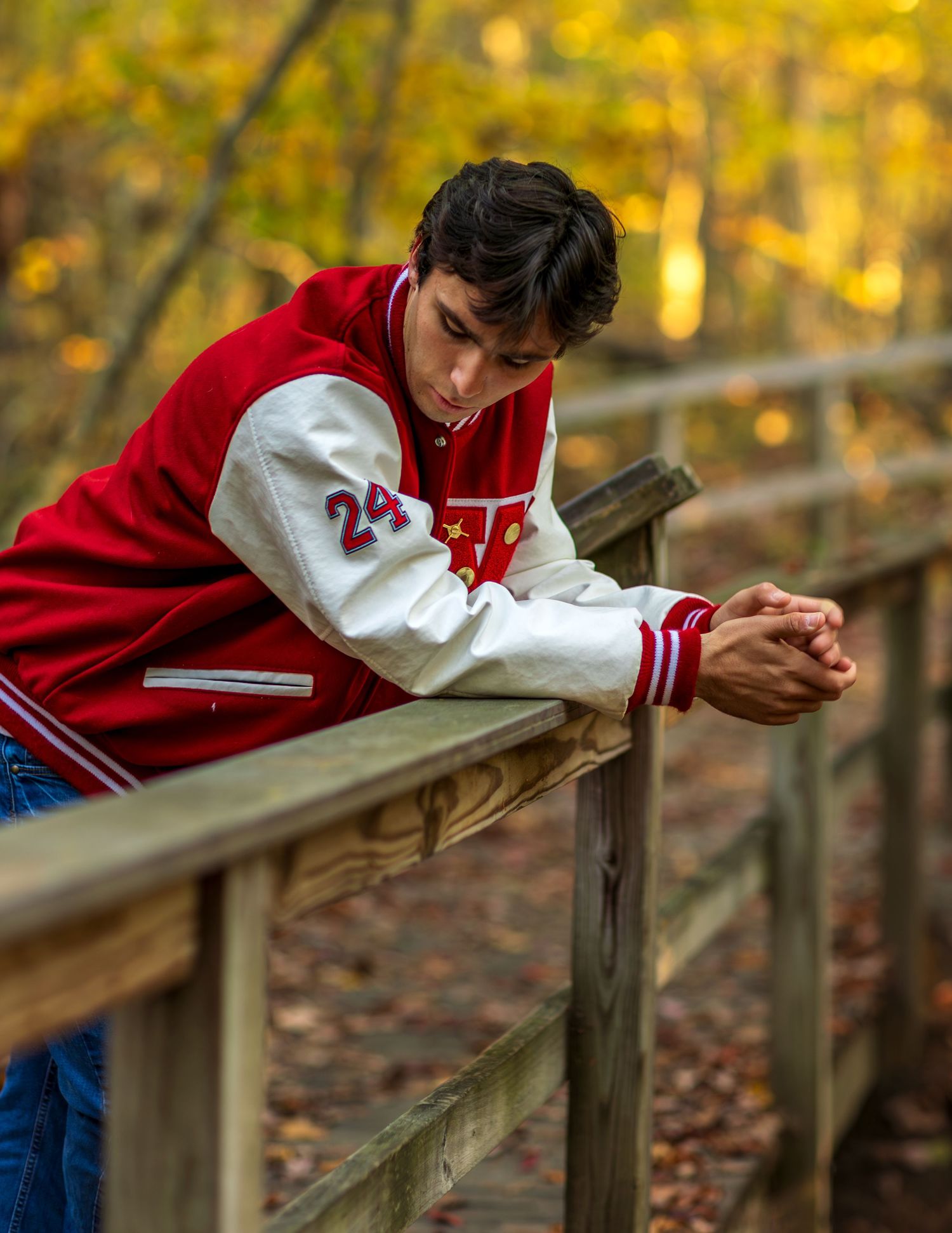 A person in a red and white varsity jacket leans thoughtfully on a wooden railing surrounded by autumn foliage.
