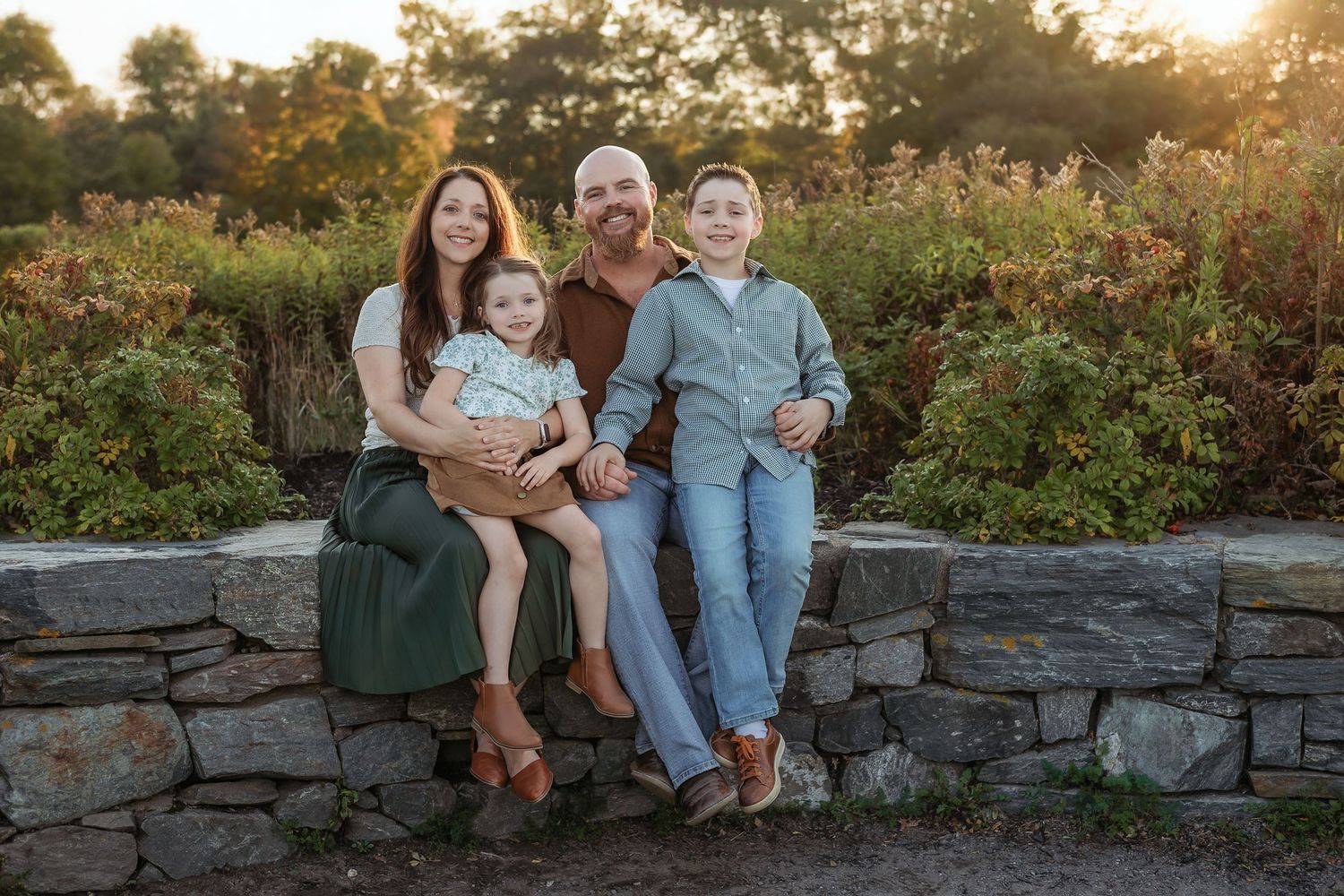 A family sits together on a stone wall at sunset in a garden setting with golden light filtering through trees.