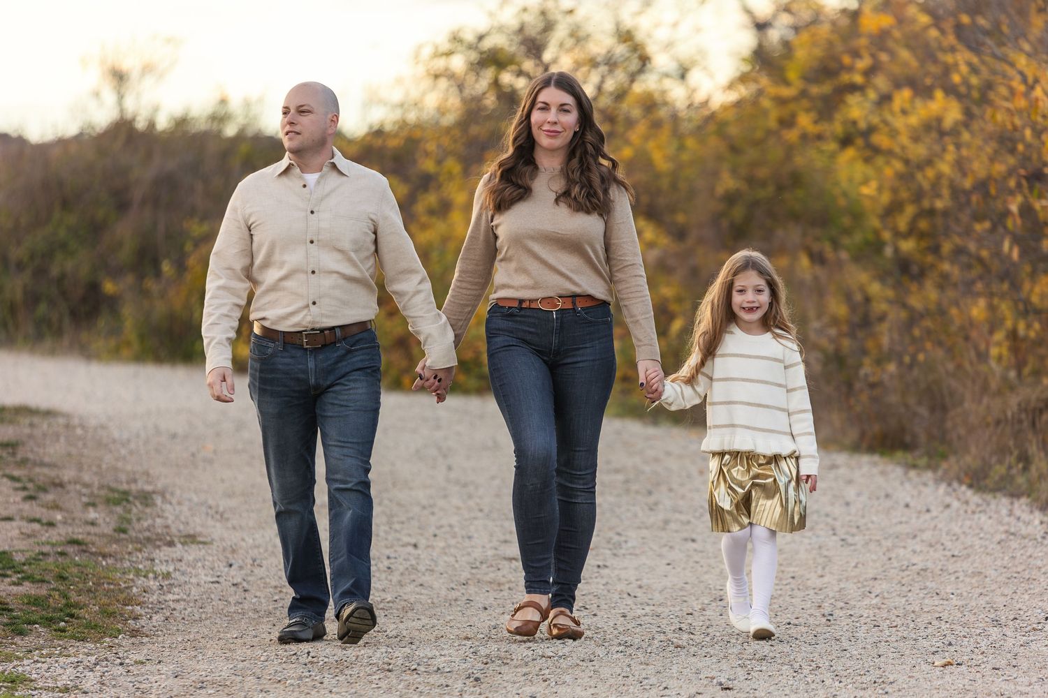 A family walks hand in hand down a gravel path during autumn with colorful fall foliage in the background.