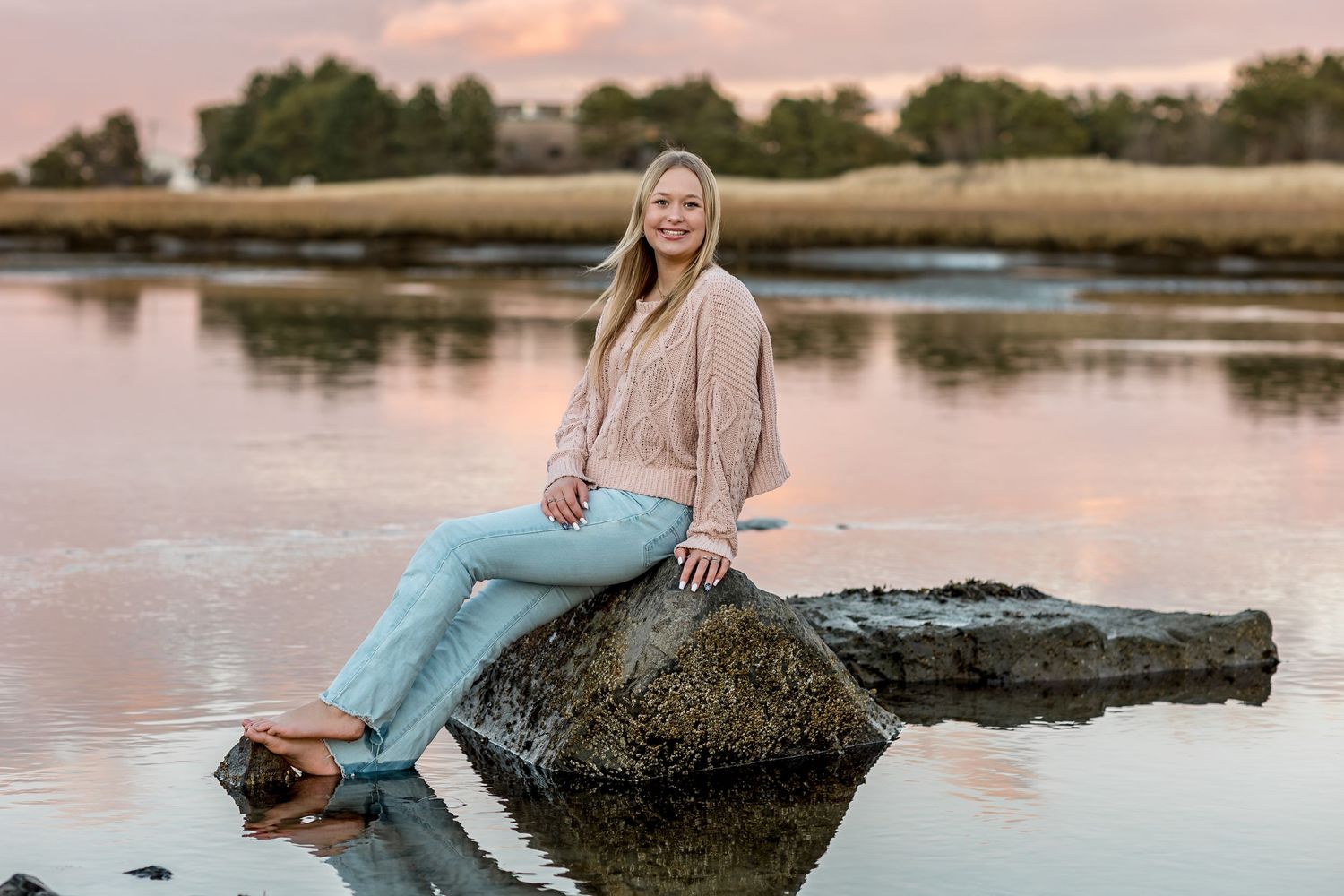 A graduating senior in beige sweater and jeans sits on rock by tranquil lake during sunset with reflection in water.