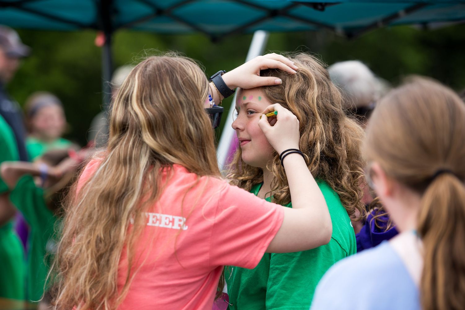 Two teenage girls in colorful t-shirts doing face painting at an outdoor youth event.