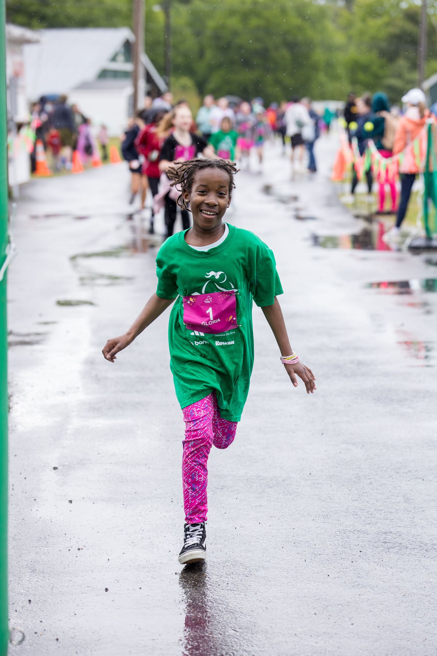 Smiling young girl in green shirt and pink leggings running on wet pavement during a kids fun run race event.