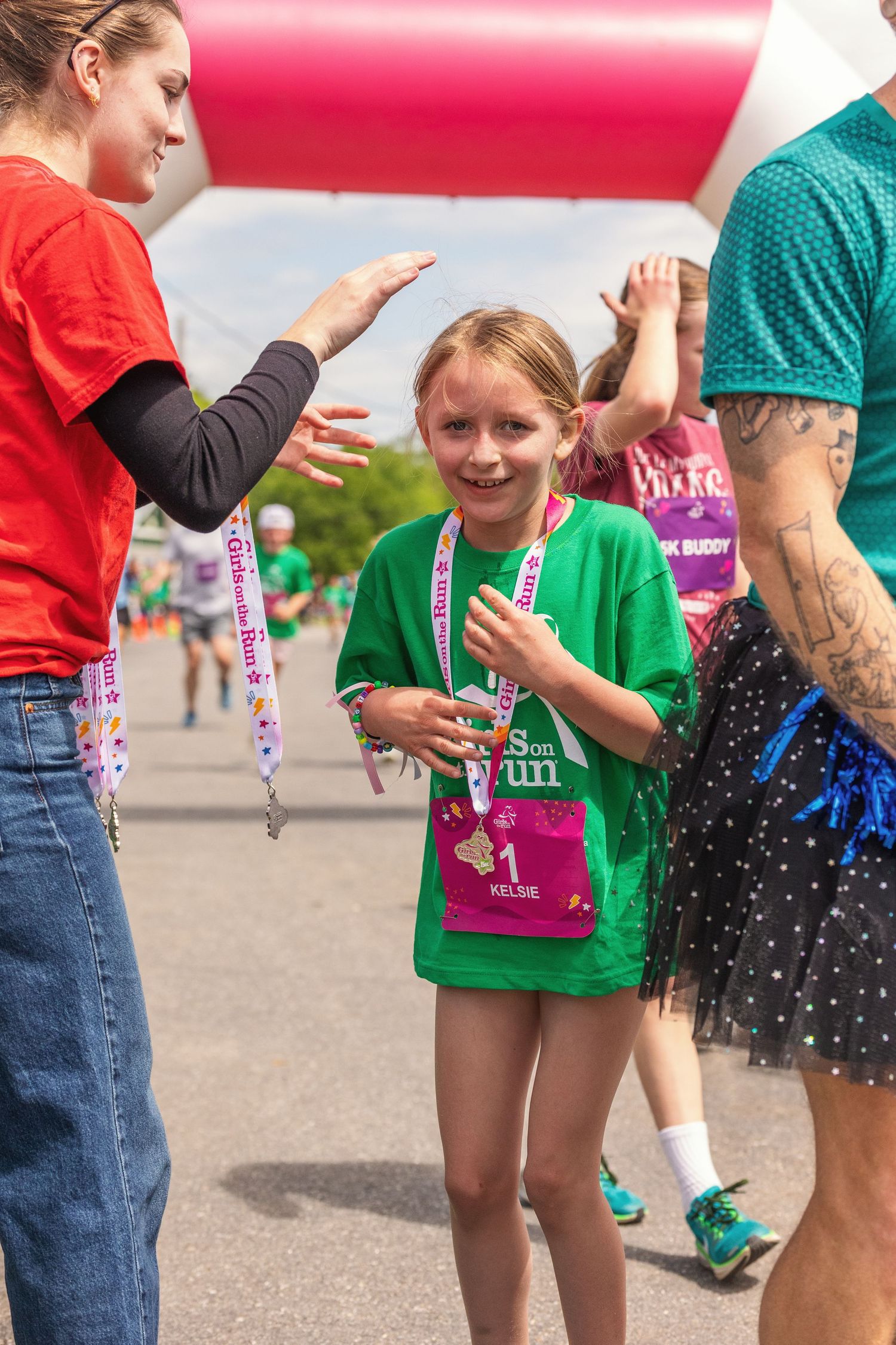 Young girl in green shirt receives high-five at finish line during kids fun run race, wearing medal and bib number 1.