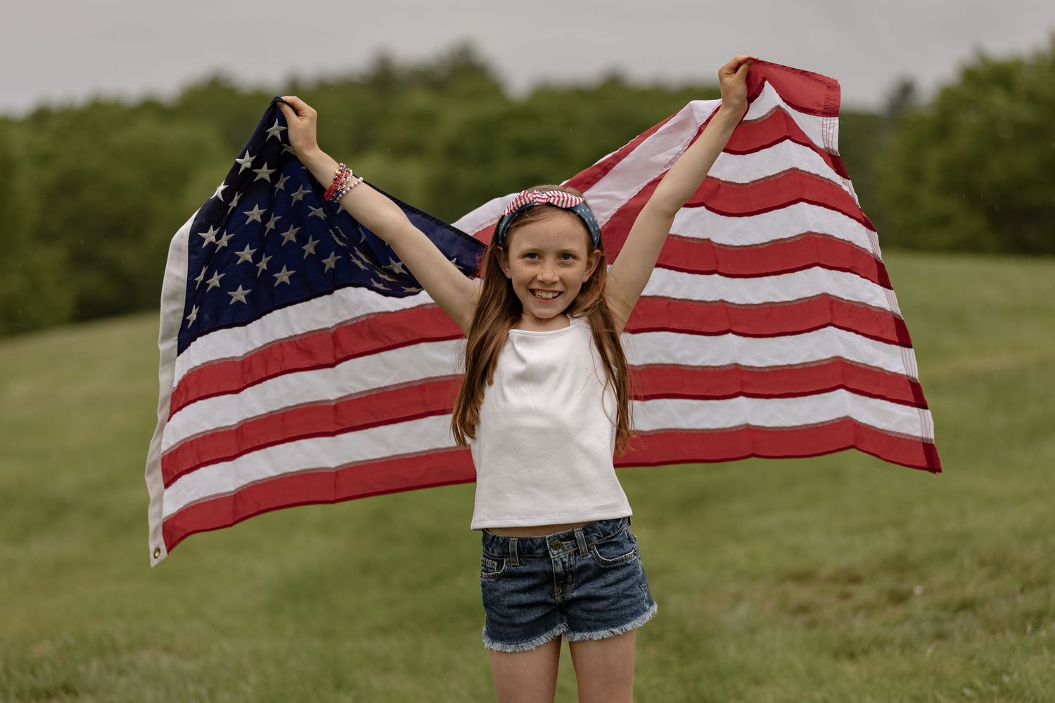 A smiling girl holds an American flag above her head in a green field, celebrating patriotism outdoors.