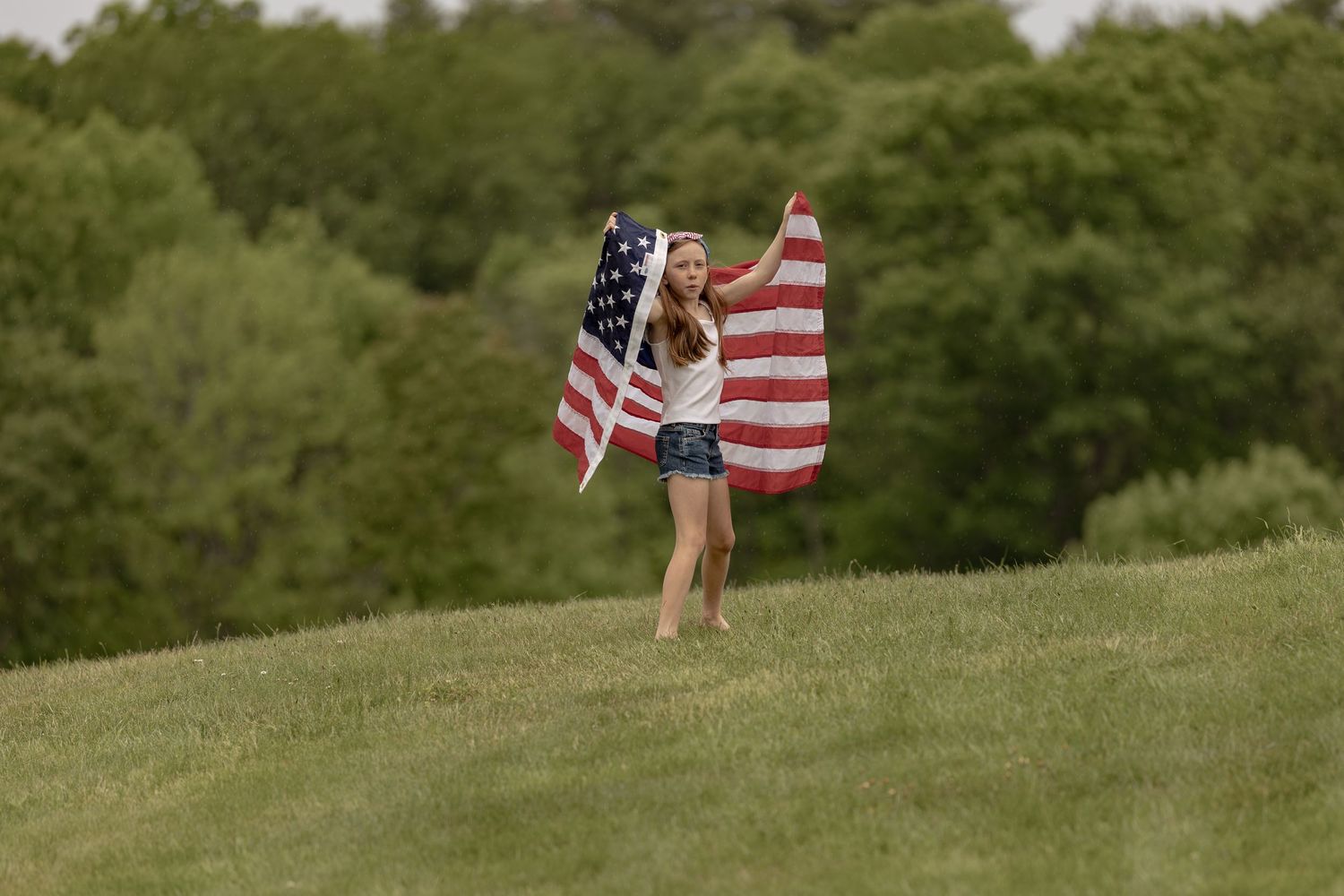 Young woman running on green hill holding large American flag outdoors.