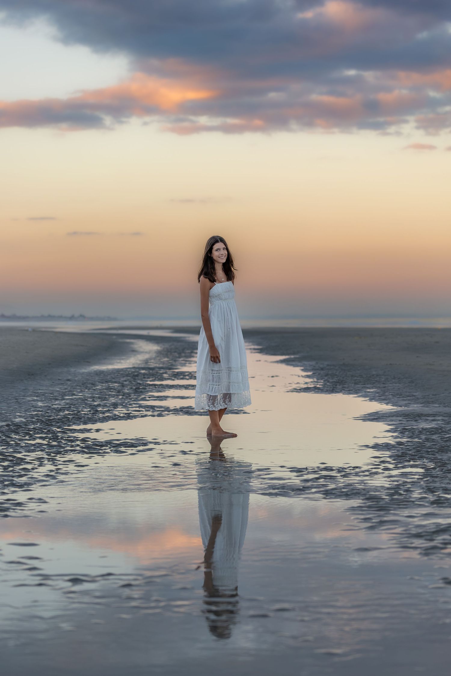 Woman in white dress stands barefoot on reflective wet beach at sunset with colorful cloudy sky.