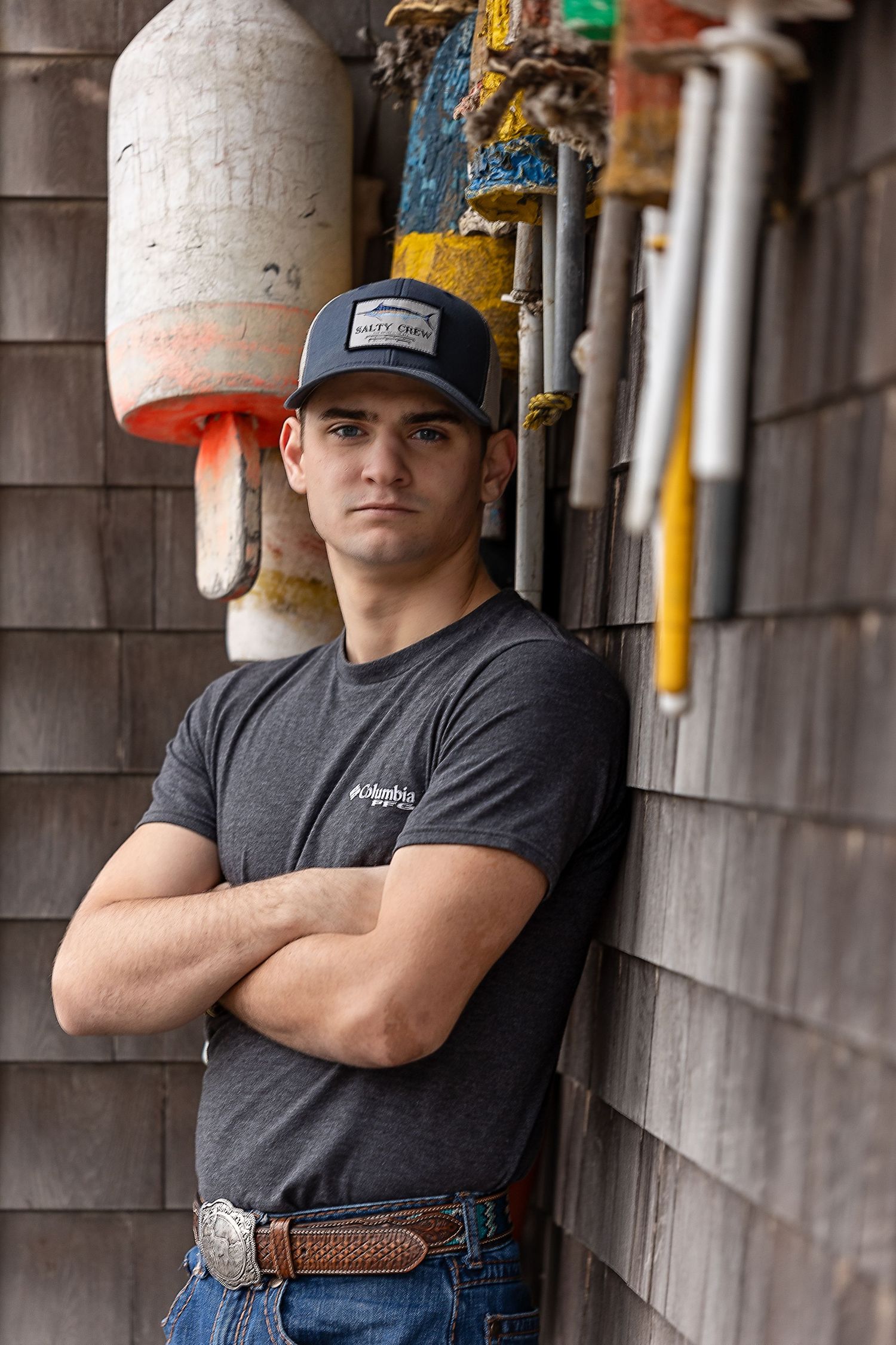 A graduating senior in dark gray t-shirt and jeans stands with arms crossed against wooden wall surrounded by buoys.