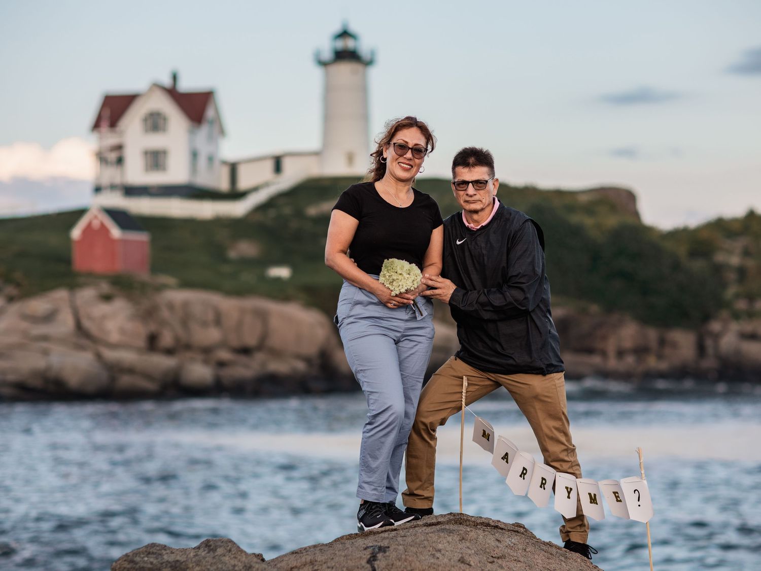 A couple sits together on coastal rocks at sunset near Nubble Lighthouse in Maine.