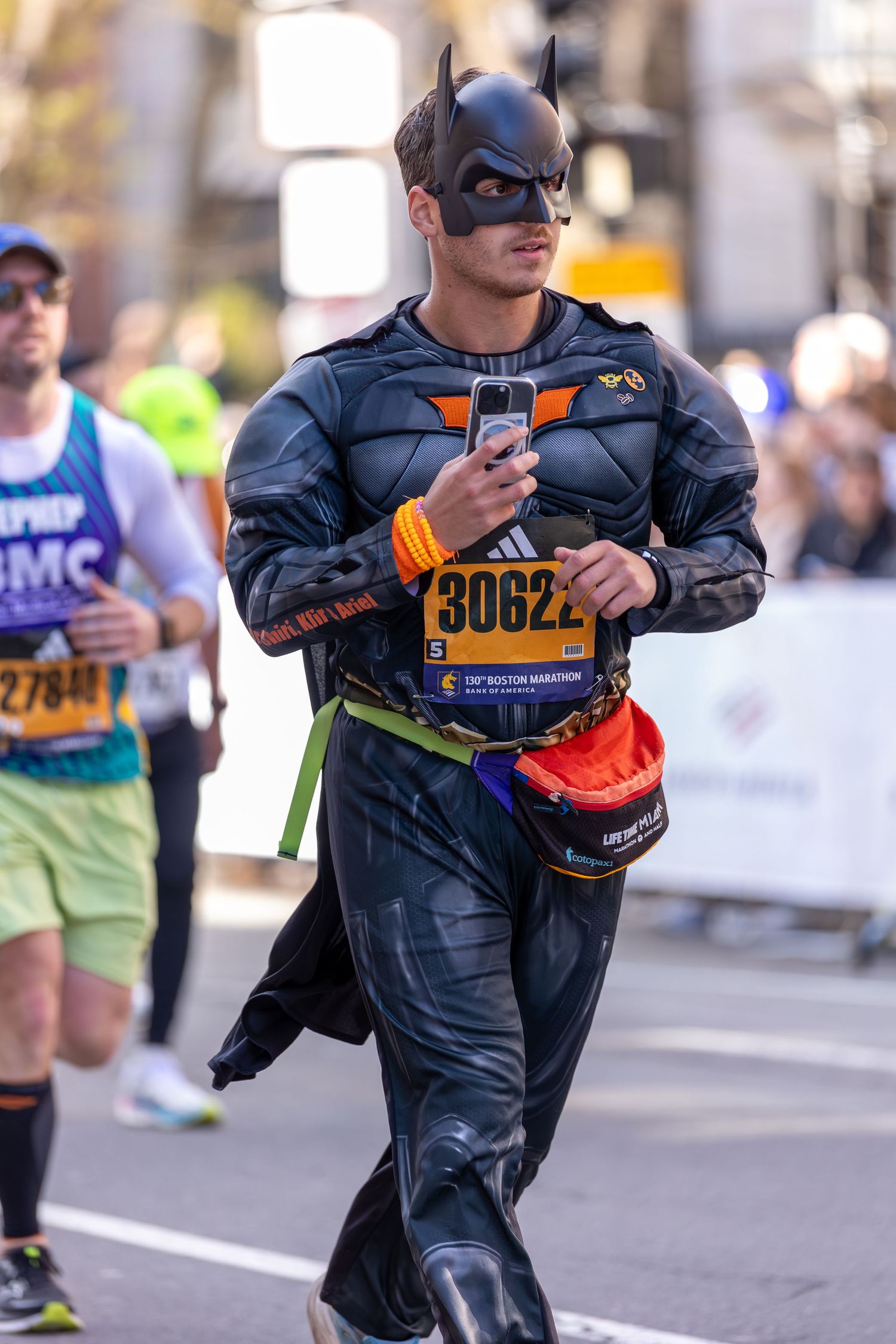 Runner dressed in Batman costume with race bib 30622 at the Boston Marathon holding a phone.