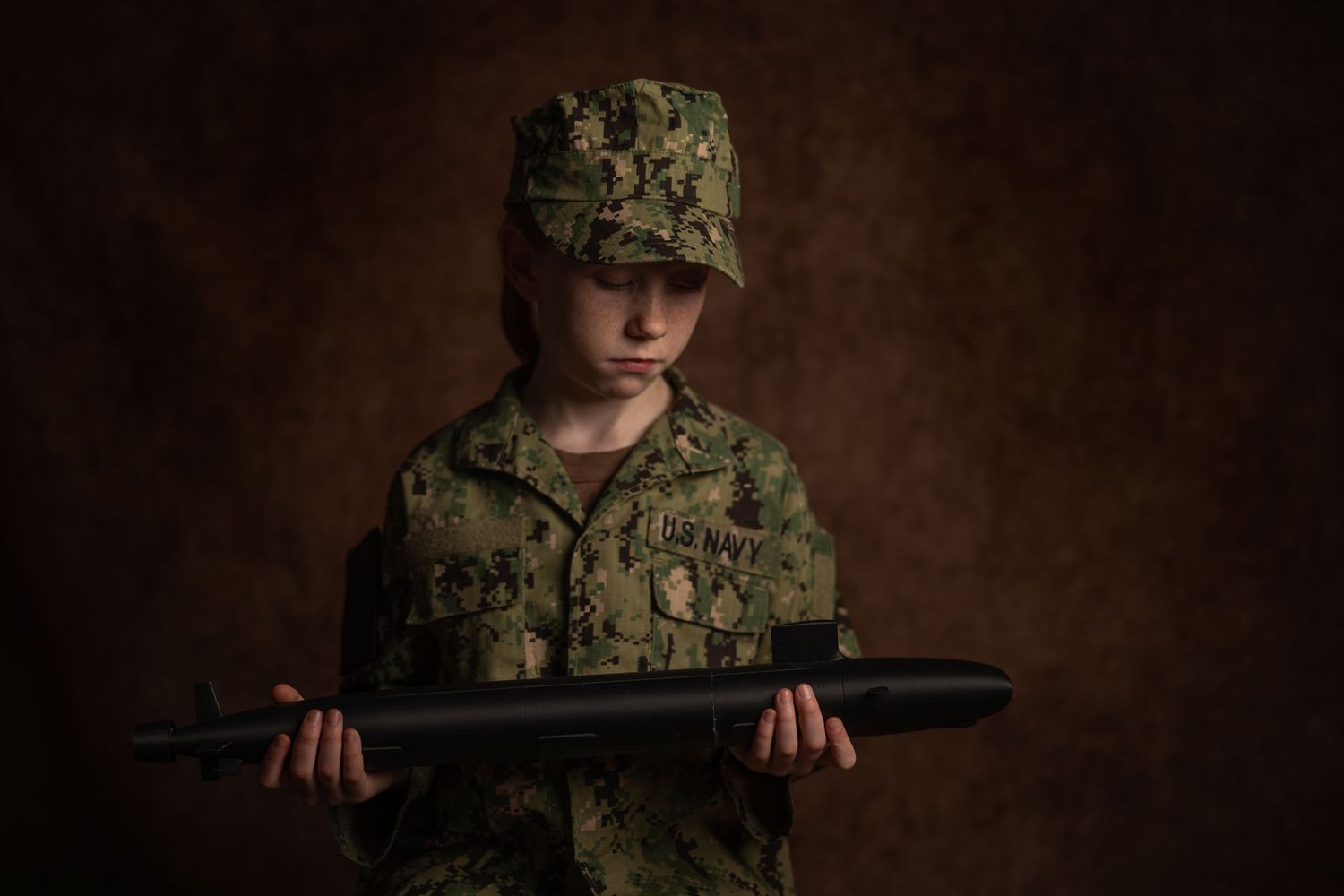 Young person in military camouflage uniform and cap holding a model submarine against a dark background.