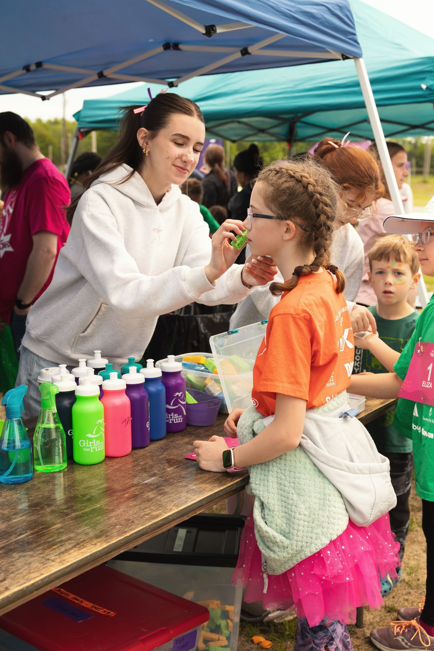 Woman painting a young girl's face at an outdoor community event booth with colorful water bottles on display.