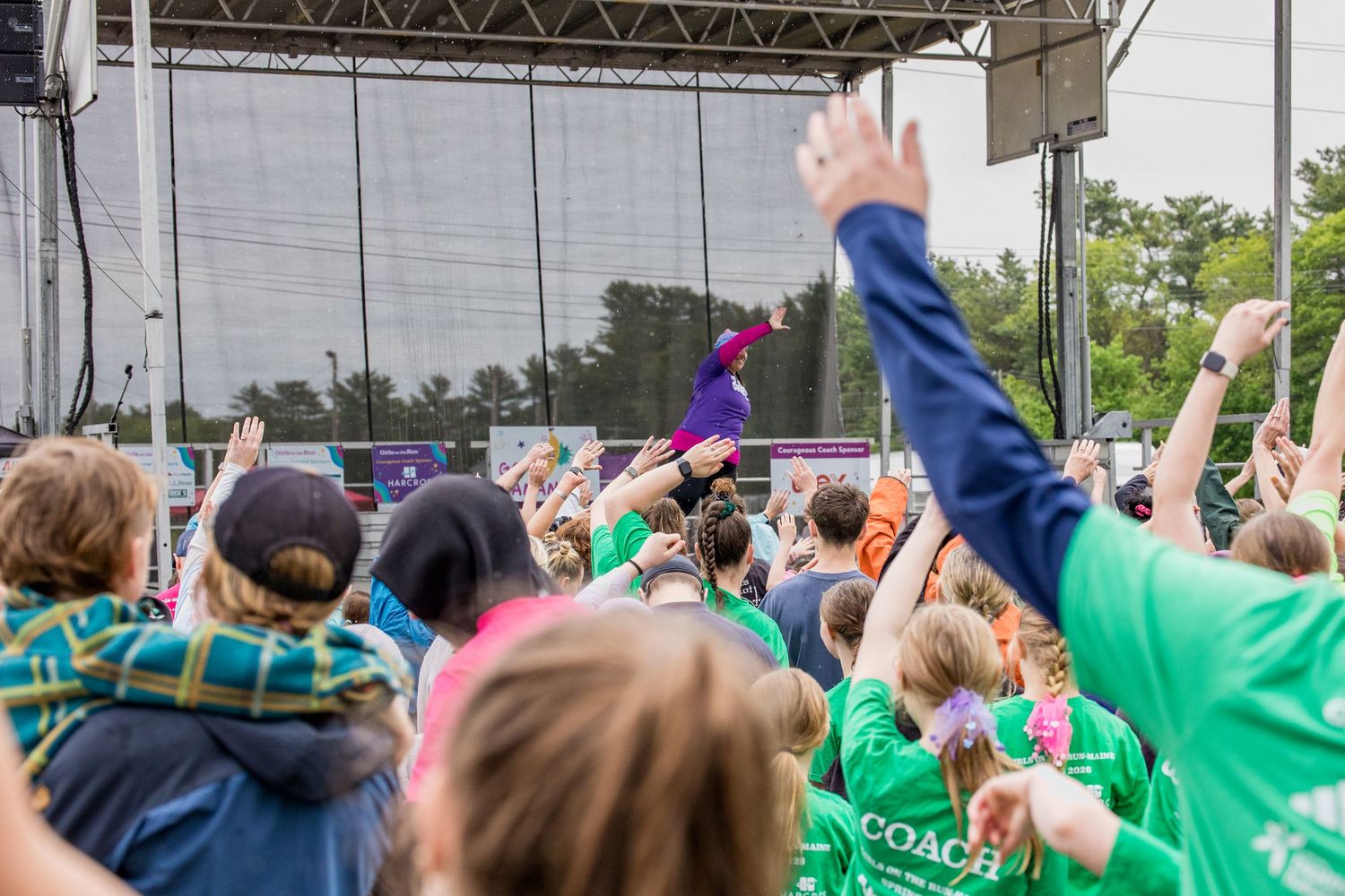 Crowd of fans raising hands at outdoor concert stage with performer in purple costume at music festival.
