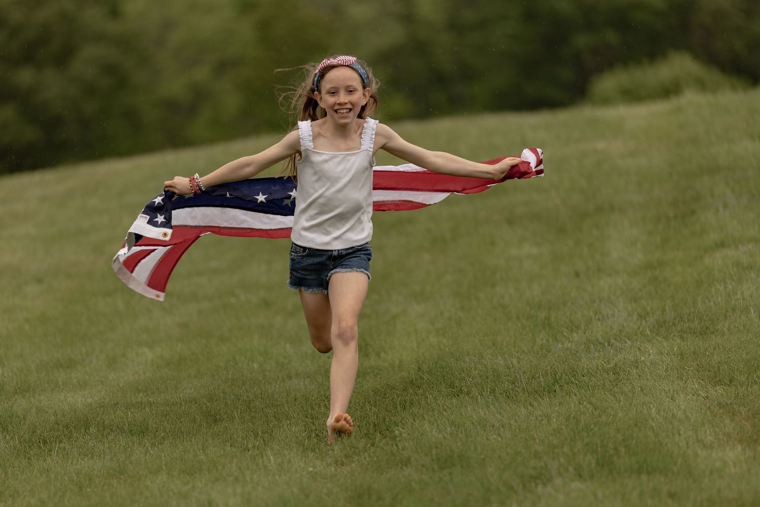 Young girl running on green grass field holding an American flag, smiling joyfully on a patriotic day.