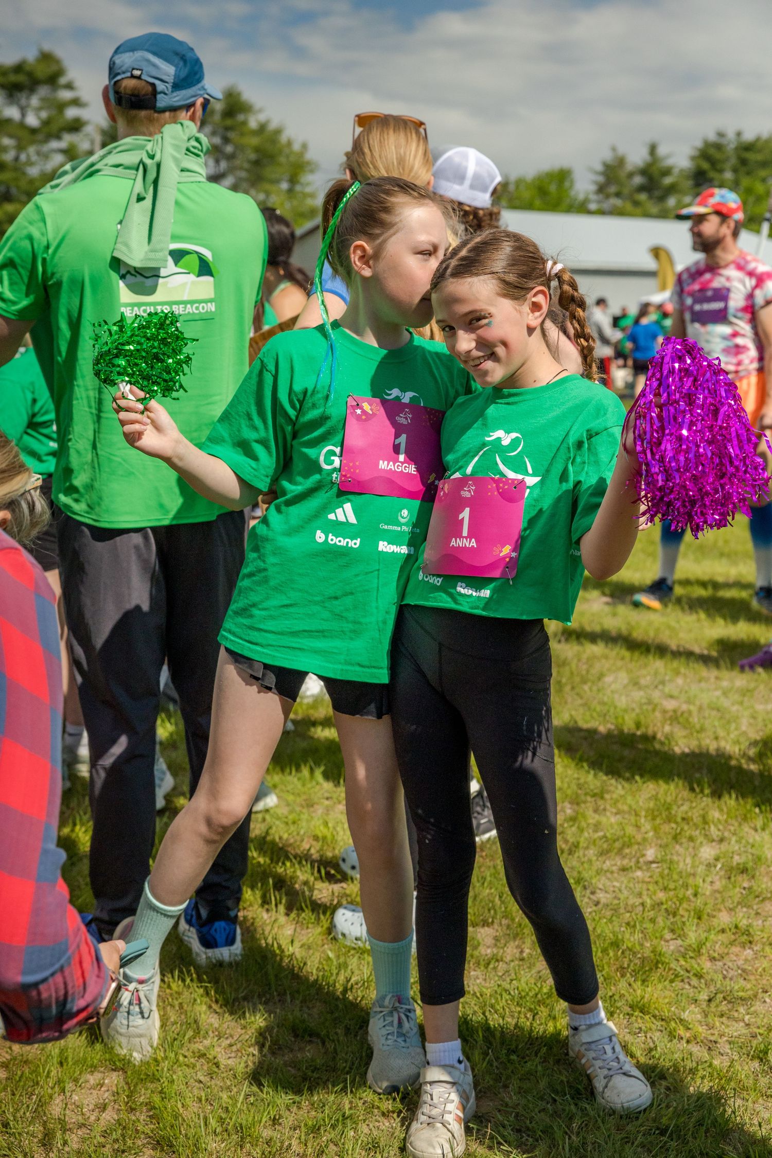 Two girls in green race shirts with pink bibs smile together at an outdoor charity fun run event.