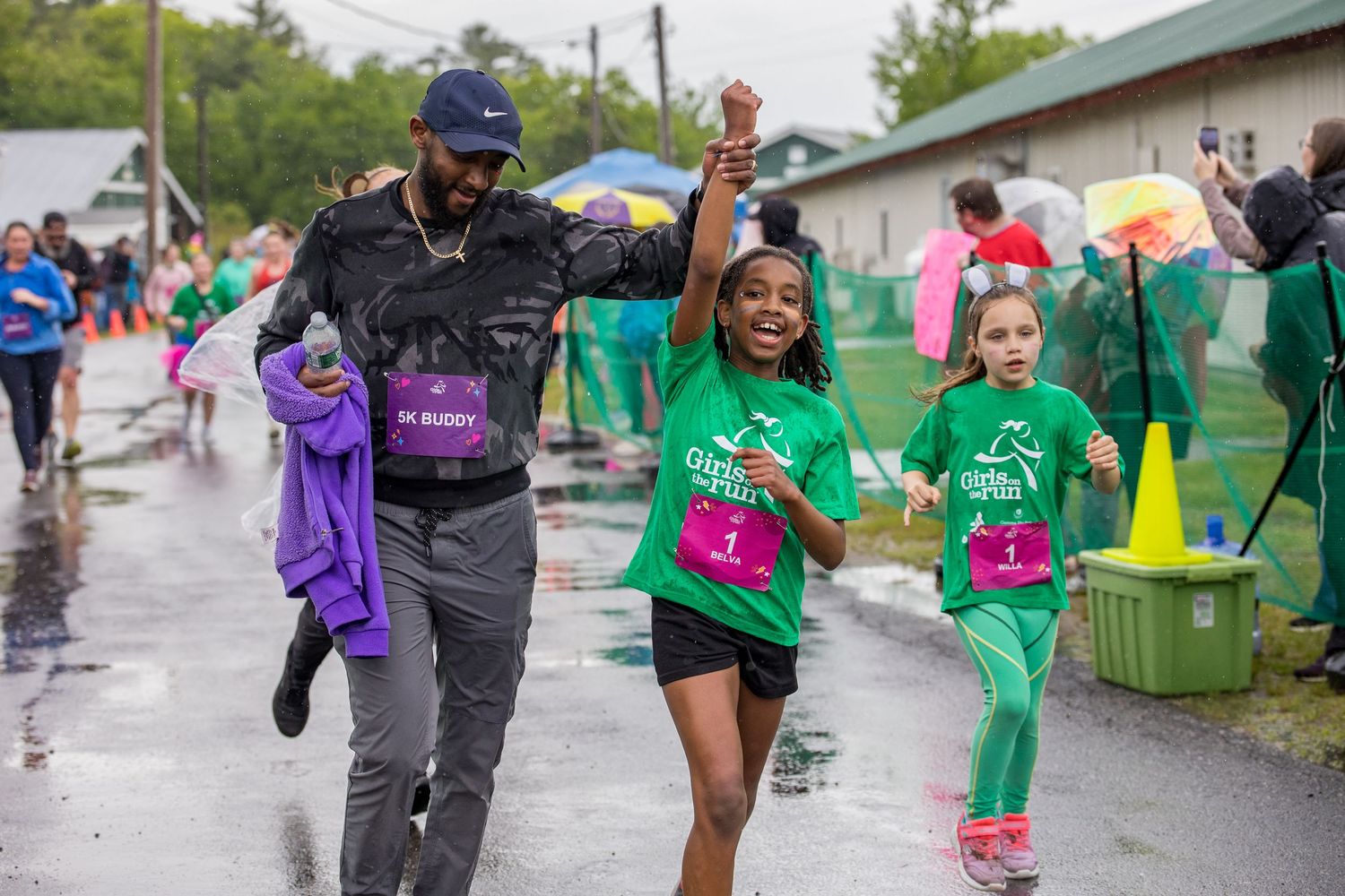 Adult and two girls in green shirts celebrate finishing a charity fun run on a rainy day.