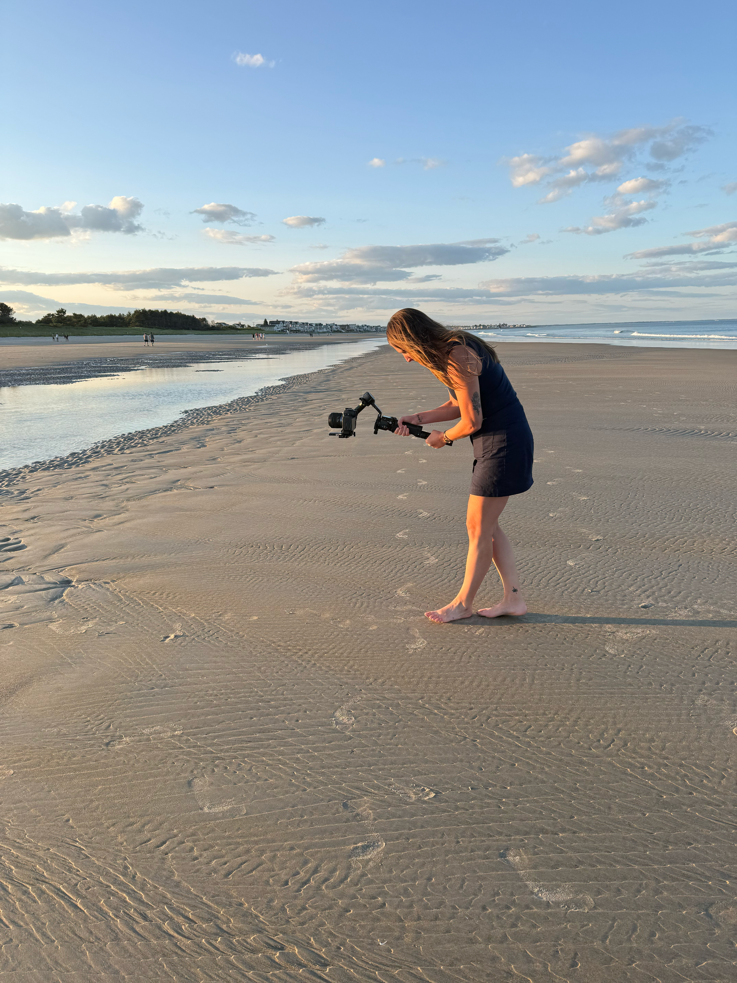 Woman filming on a sandy beach at golden hour using a camera gimbal stabilizer with calm ocean waves in the background.