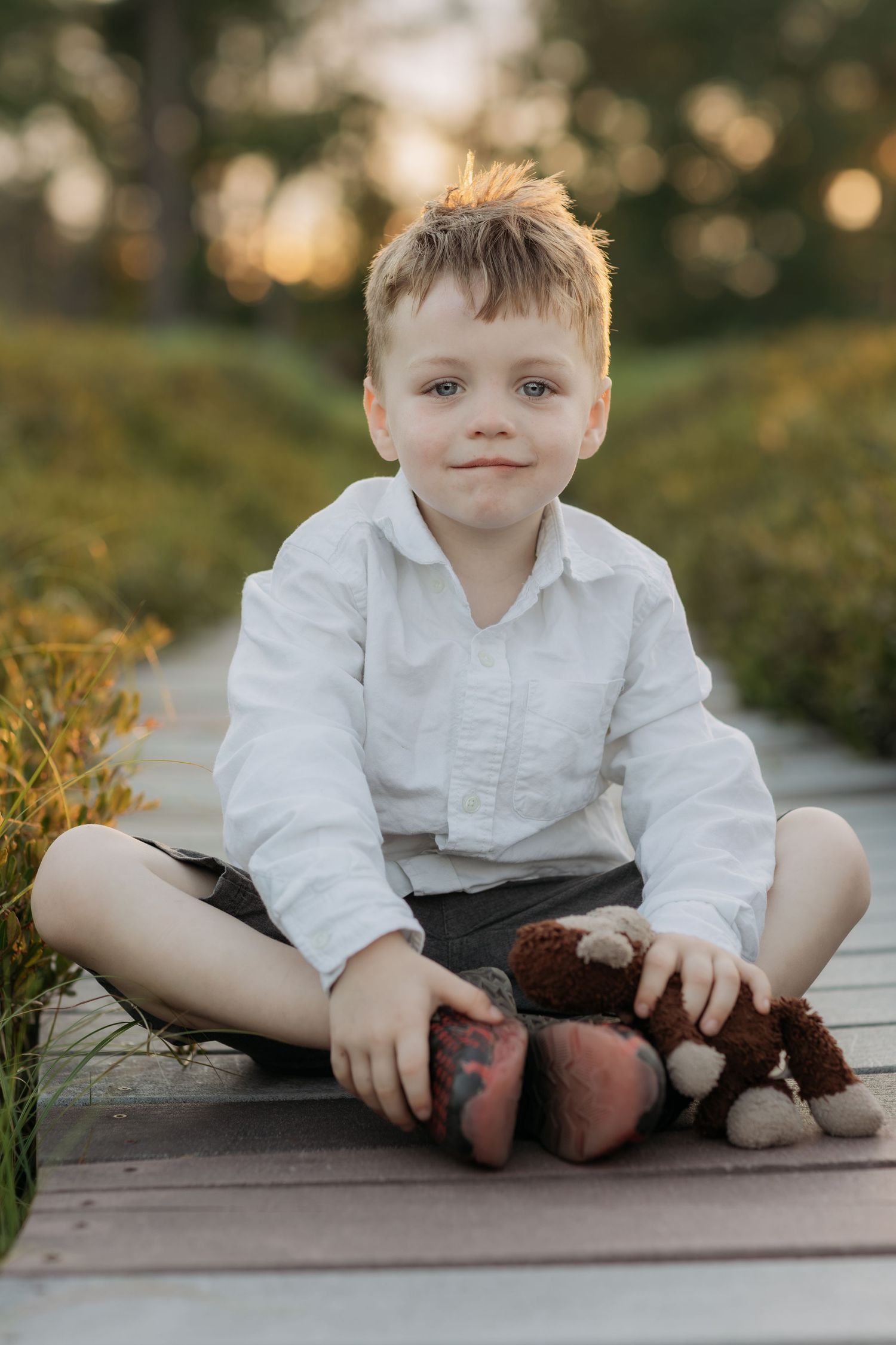 Young boy in white shirt sitting cross-legged on wooden path holding a brown teddy bear at sunset.