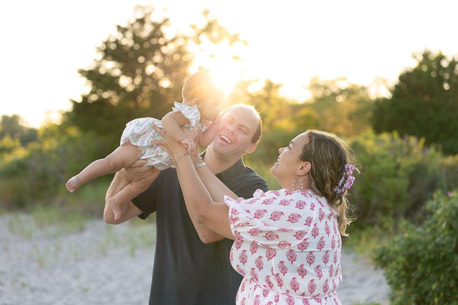 Happy family with parents lifting baby at golden hour beach, sunlight glowing through trees in background.