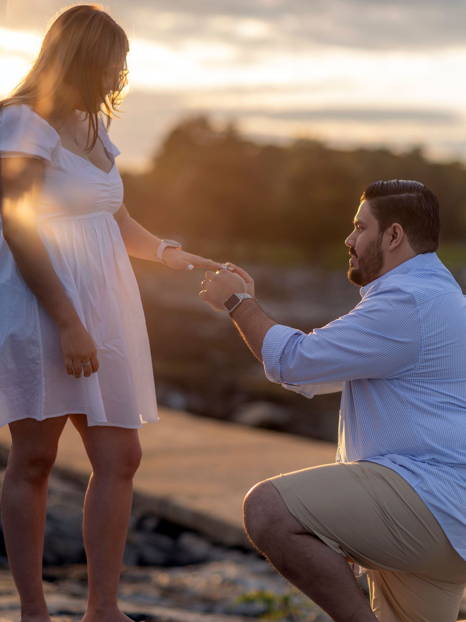 Man kneeling on one knee proposing to woman in white dress at sunset on rocky shore.